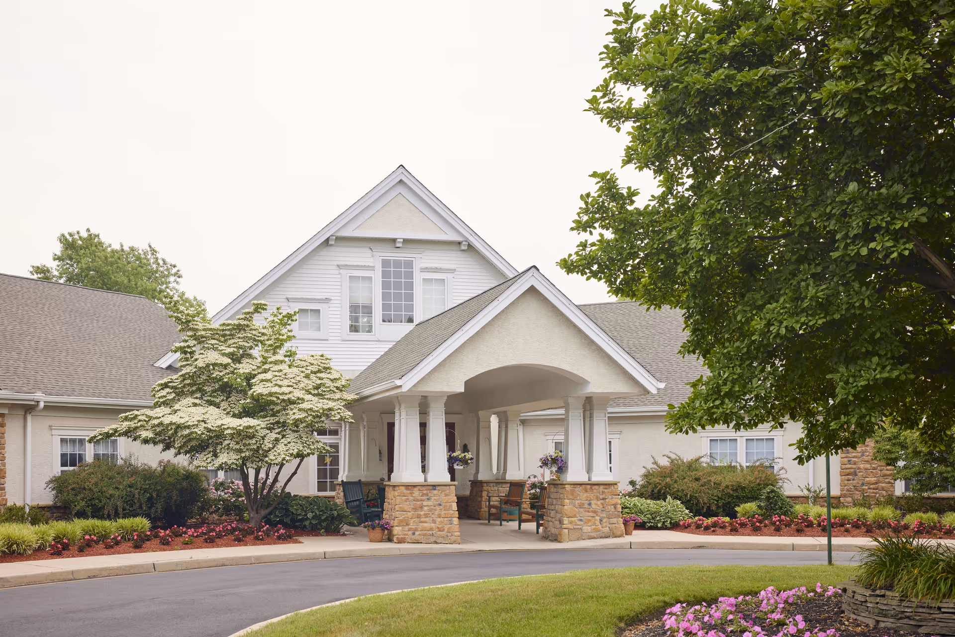 Front exterior view of a senior living facility building with a covered entrance supported by white columns on stone bases. The building is surrounded by well-maintained landscaping including flowering plants, bushes, and trees. There are chairs under the covered entrance area.