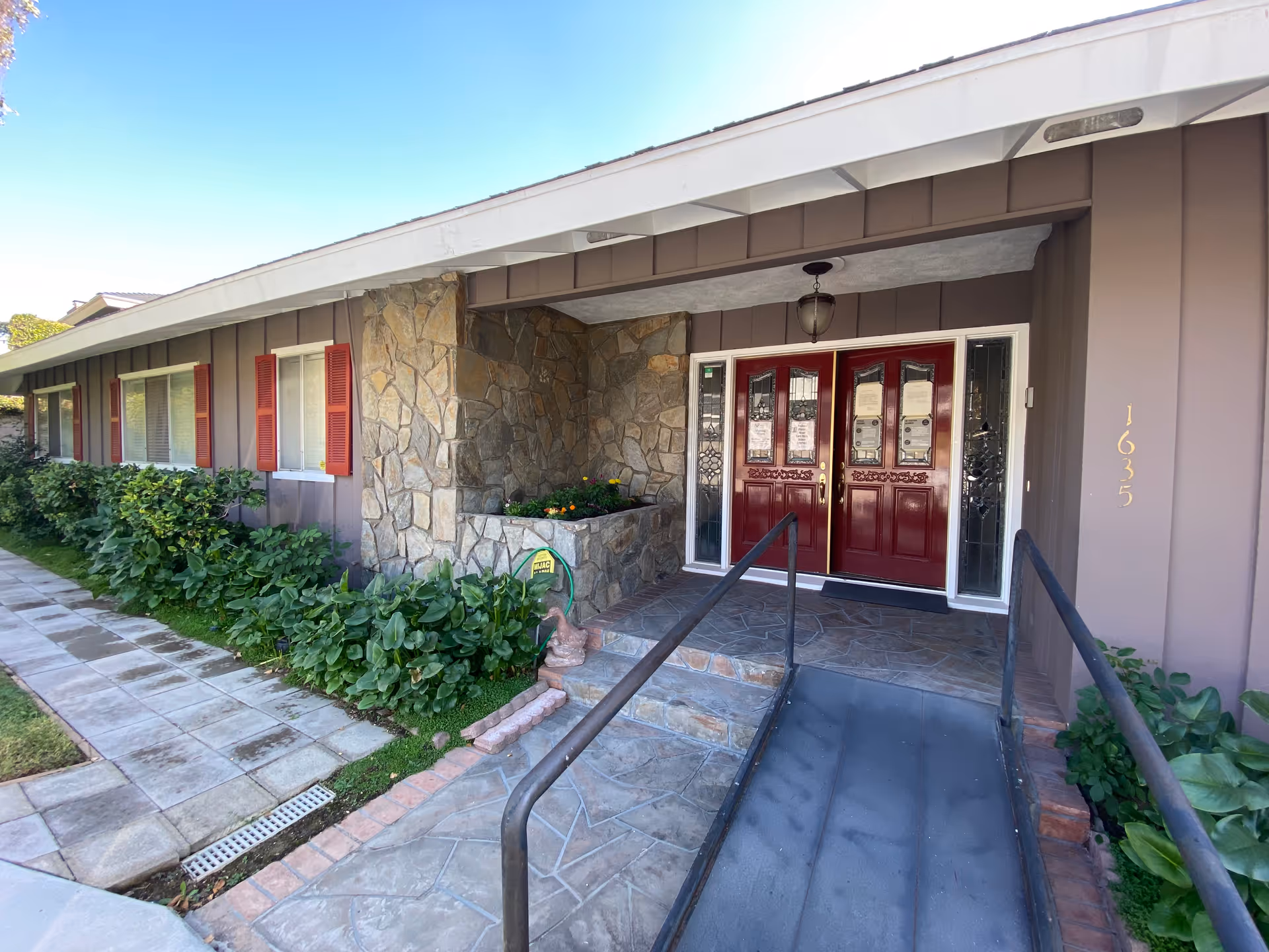 Front entrance of a single-story senior living facility with a stone facade, double red doors and an access ramp.