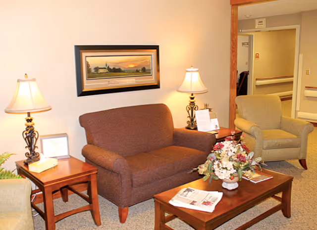 A cozy sitting area in a senior living facility with a brown loveseat, a beige armchair, two wooden side tables each with a lamp, and a wooden coffee table with a floral arrangement and newspapers. A framed landscape painting hangs on the wall above the loveseat. The background shows a hallway with room number 200 visible.