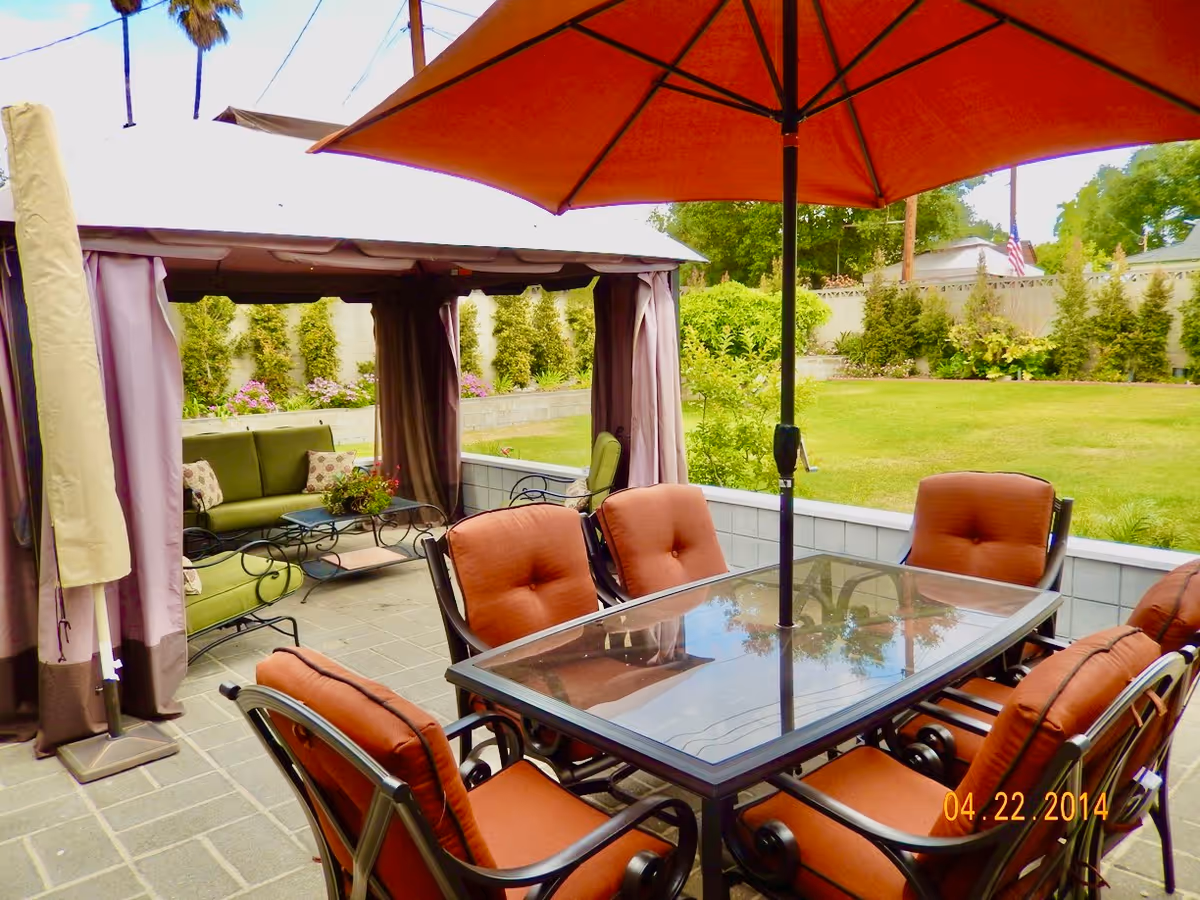 Outdoor patio area with a glass-top table surrounded by six cushioned chairs under a large orange umbrella. In the background, there is a gazebo with green cushioned seating and a small table, set on a tiled floor with a grassy yard and trees beyond a low wall.