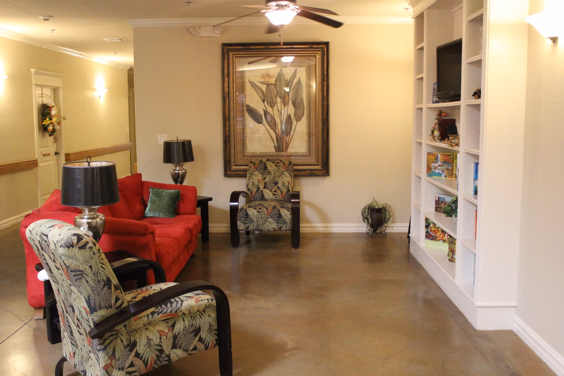 A well-lit communal living room with patterned armchairs, a red sofa, side lamps, a framed botanical print, and built-in shelving.