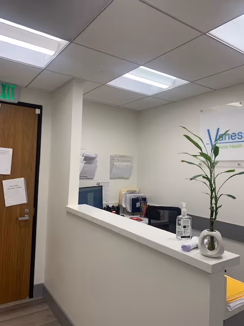 Reception counter inside Vanessa Home Health Services, plant on desk, sanitizer bottle, doorway to left.