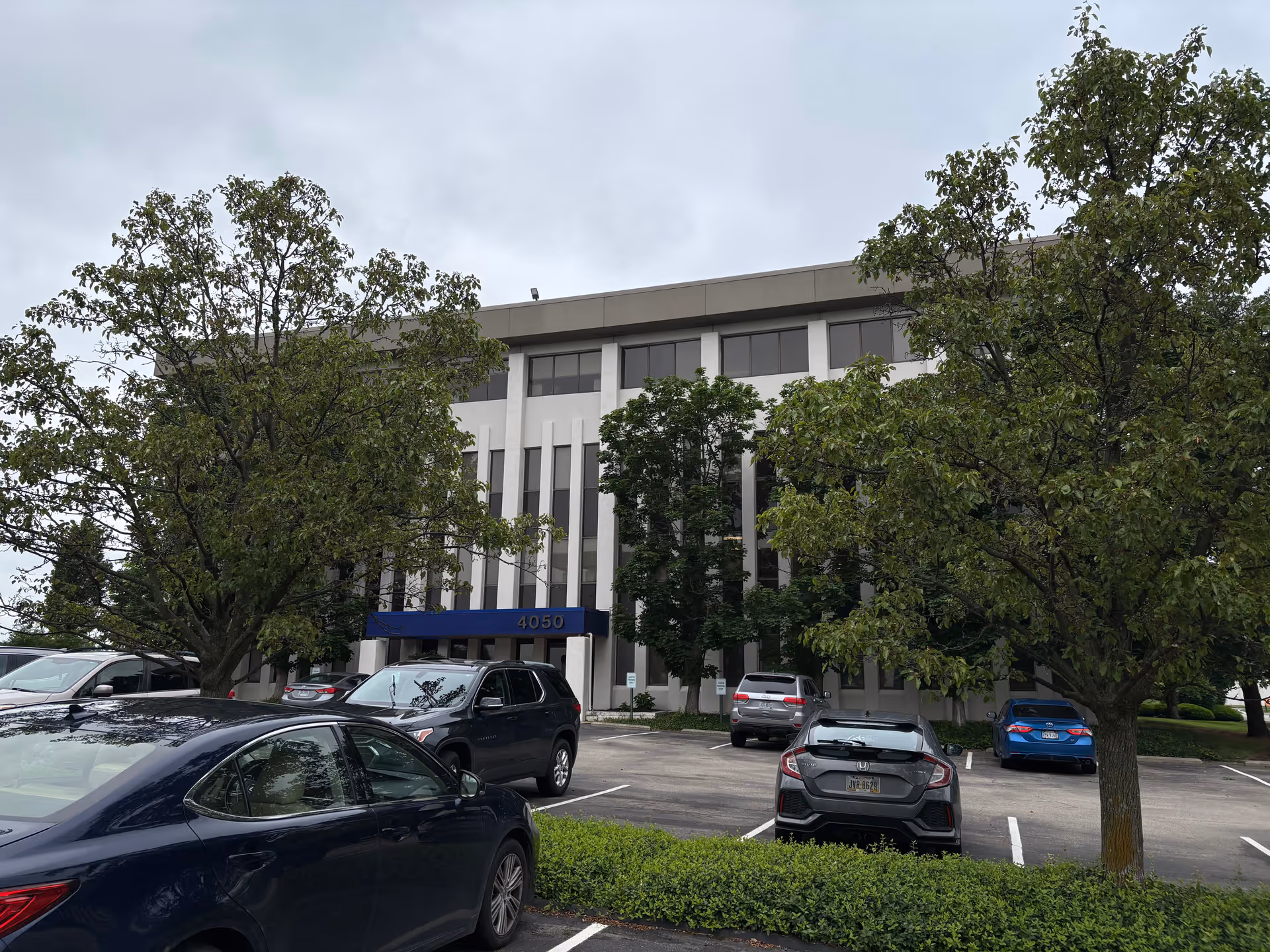 Front view of a multi-story care facility with a blue entrance canopy and parked cars.