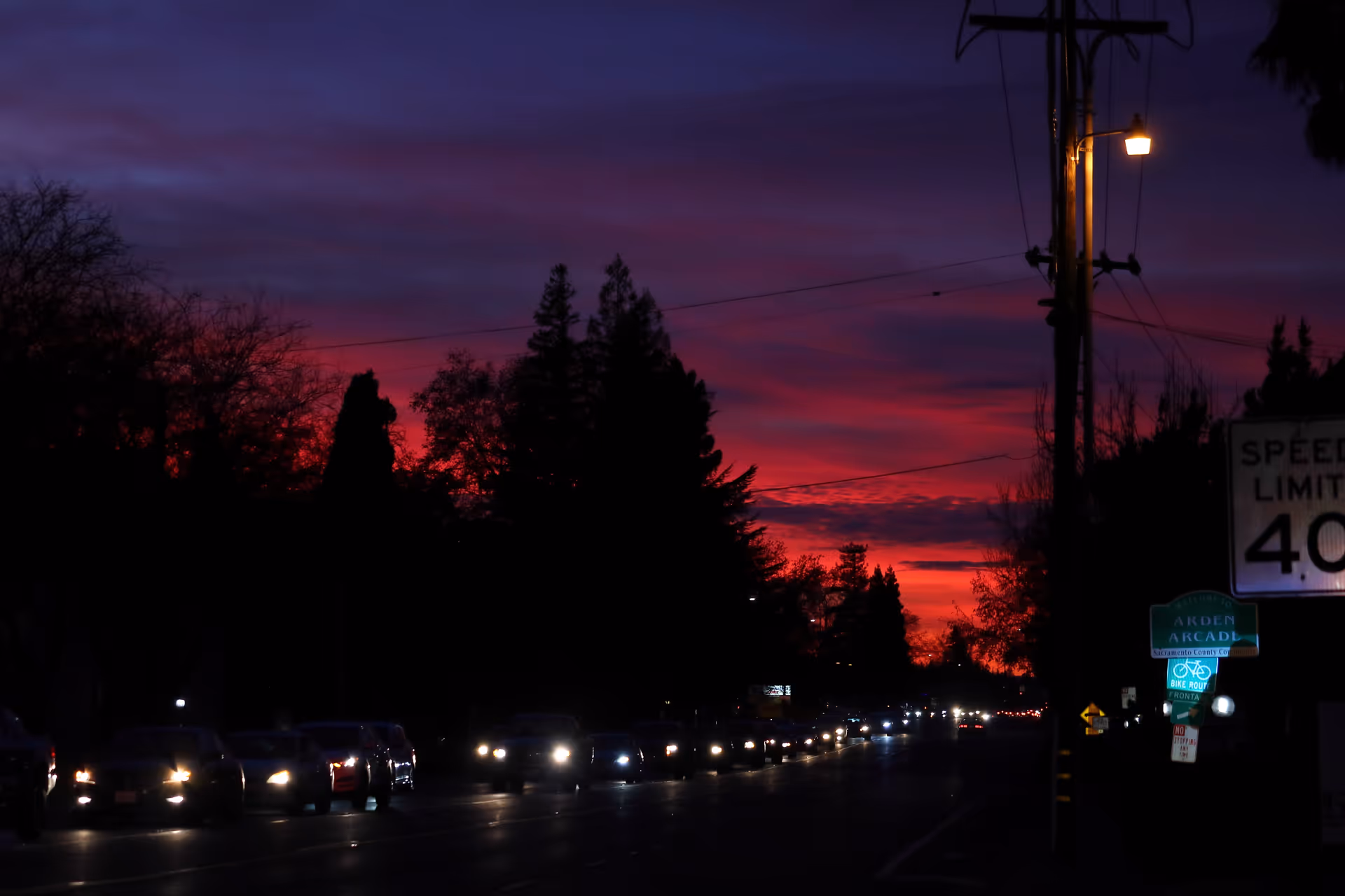 A dark street scene at sunset with cars along the road and power lines.