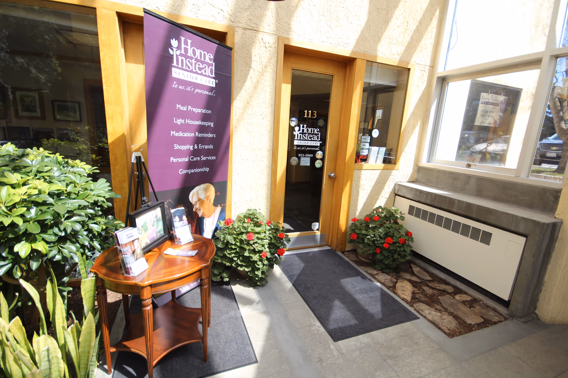 Entrance of a Home Instead facility with a wooden door, banner, and potted plants.