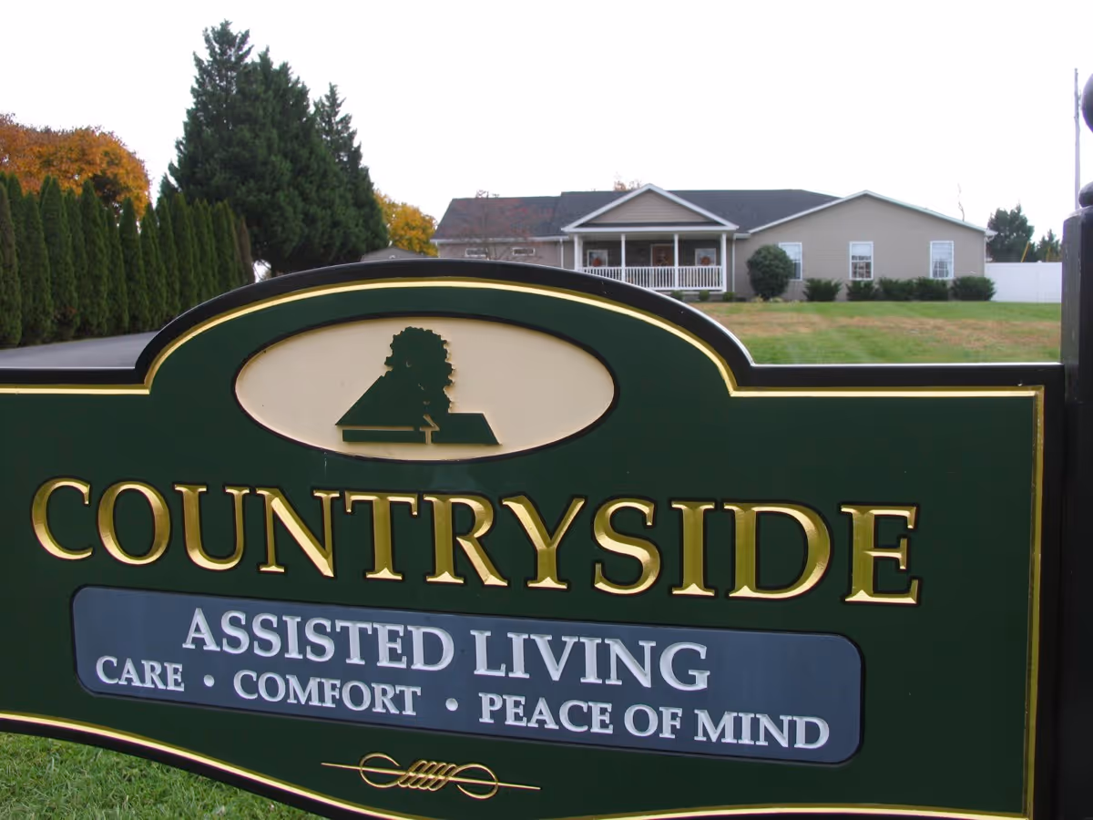 A green and gold sign for Countryside Assisted Living with the words 'Care, Comfort, Peace of Mind' in front of a single-story building with a porch and a lawn.