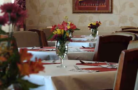 Dining room with tables set for meals, floral centerpieces, and empty chairs.