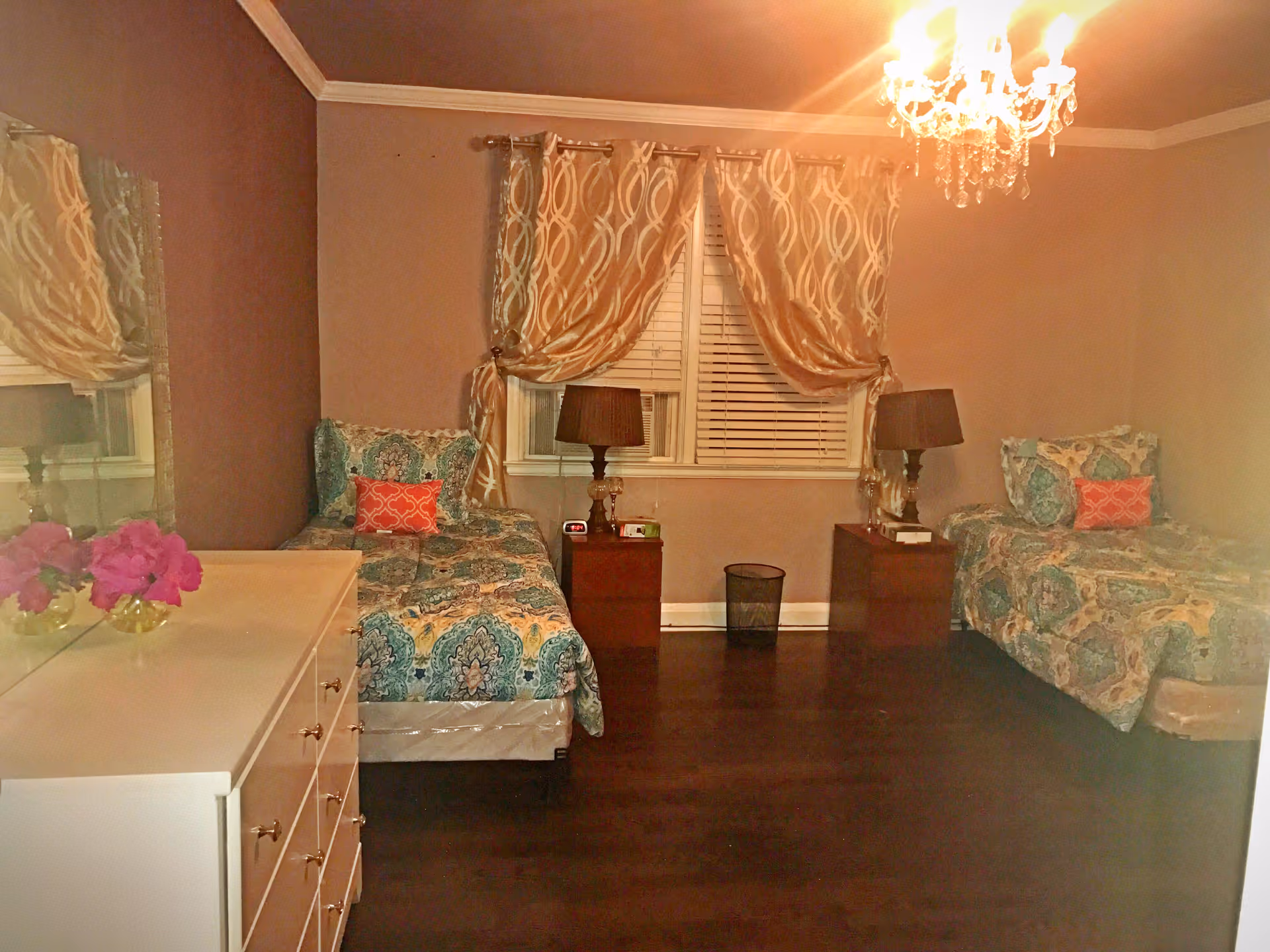 Two patterned twin beds flank a window with curtains, a chandelier, nightstands, and a dresser.