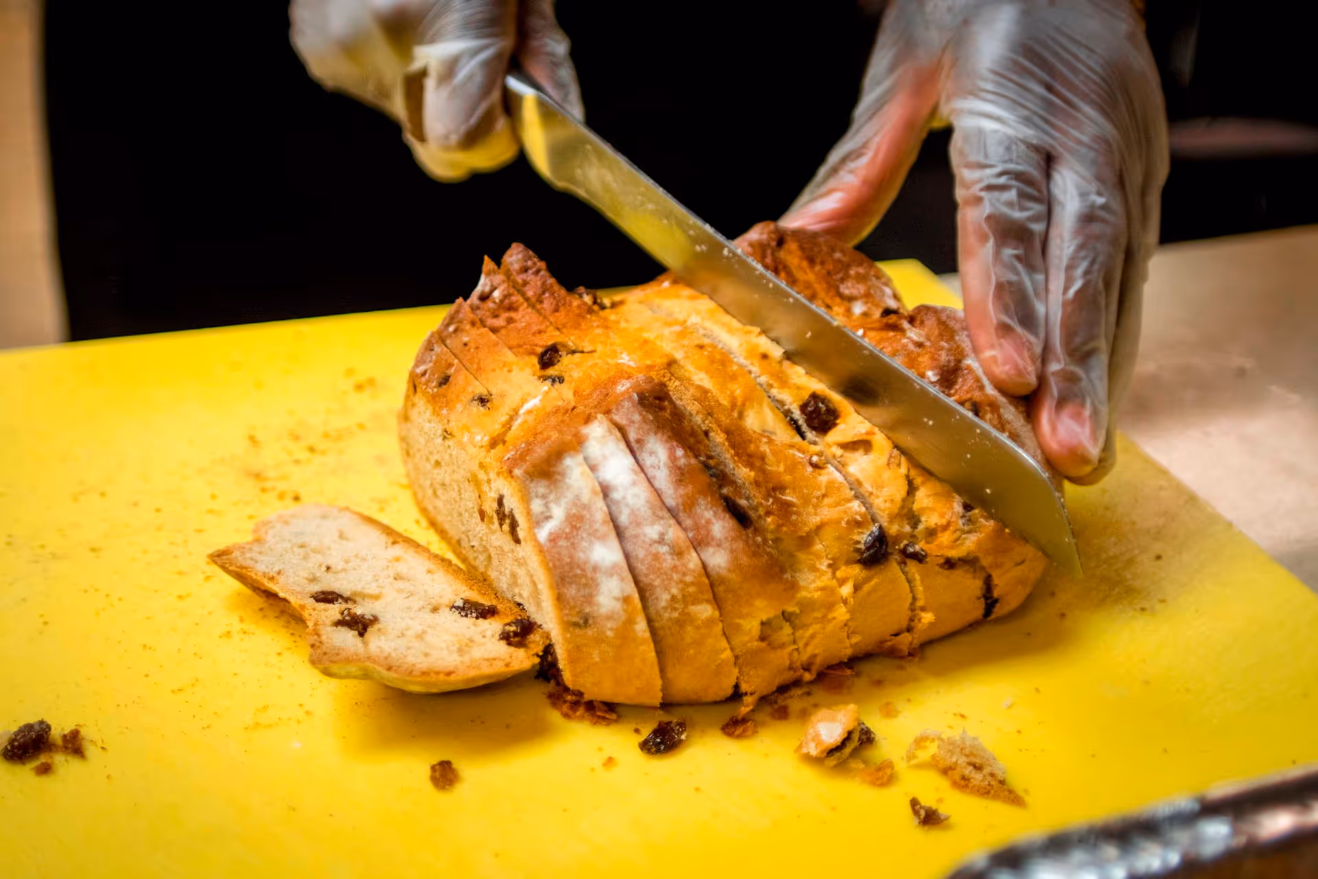 Gloved hands slice a loaf of bread on a yellow cutting board in a kitchen.