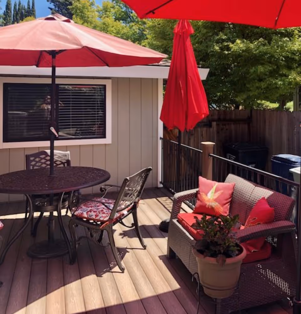 Outdoor patio area with a round metal table and two chairs with floral cushions, a wicker loveseat with red cushions and decorative pillows, two red umbrellas providing shade, a potted plant on the floor, and a wooden fence with trees in the background.