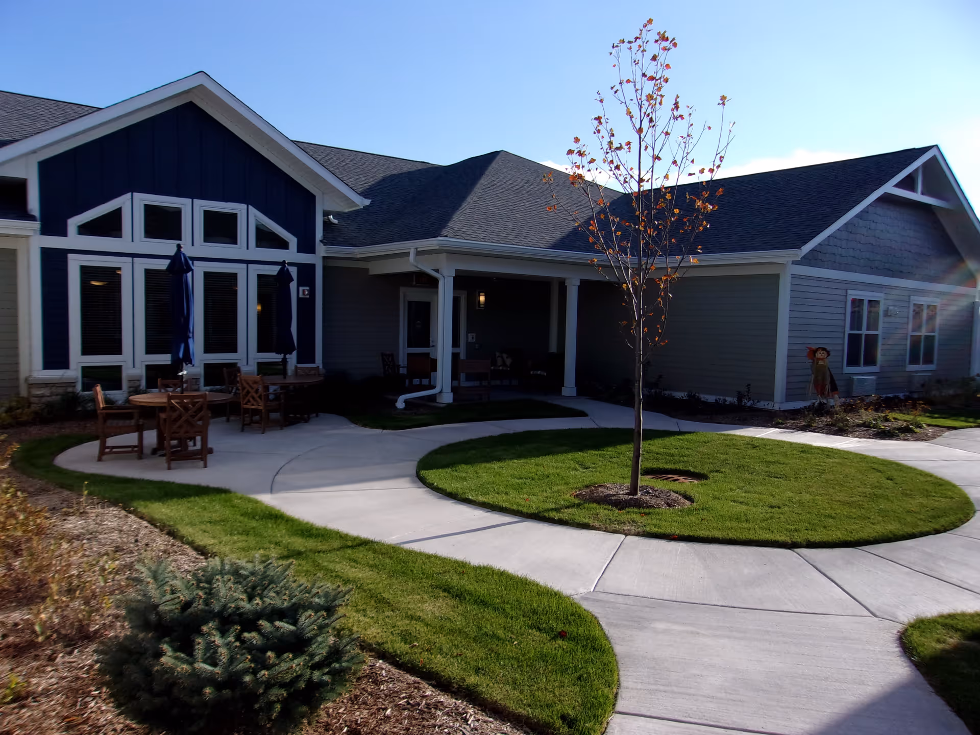 Exterior view of a senior living facility building with a circular concrete walkway surrounding a small tree and green lawn. There are outdoor tables and chairs with closed umbrellas near the building, which has large windows and a covered porch area.