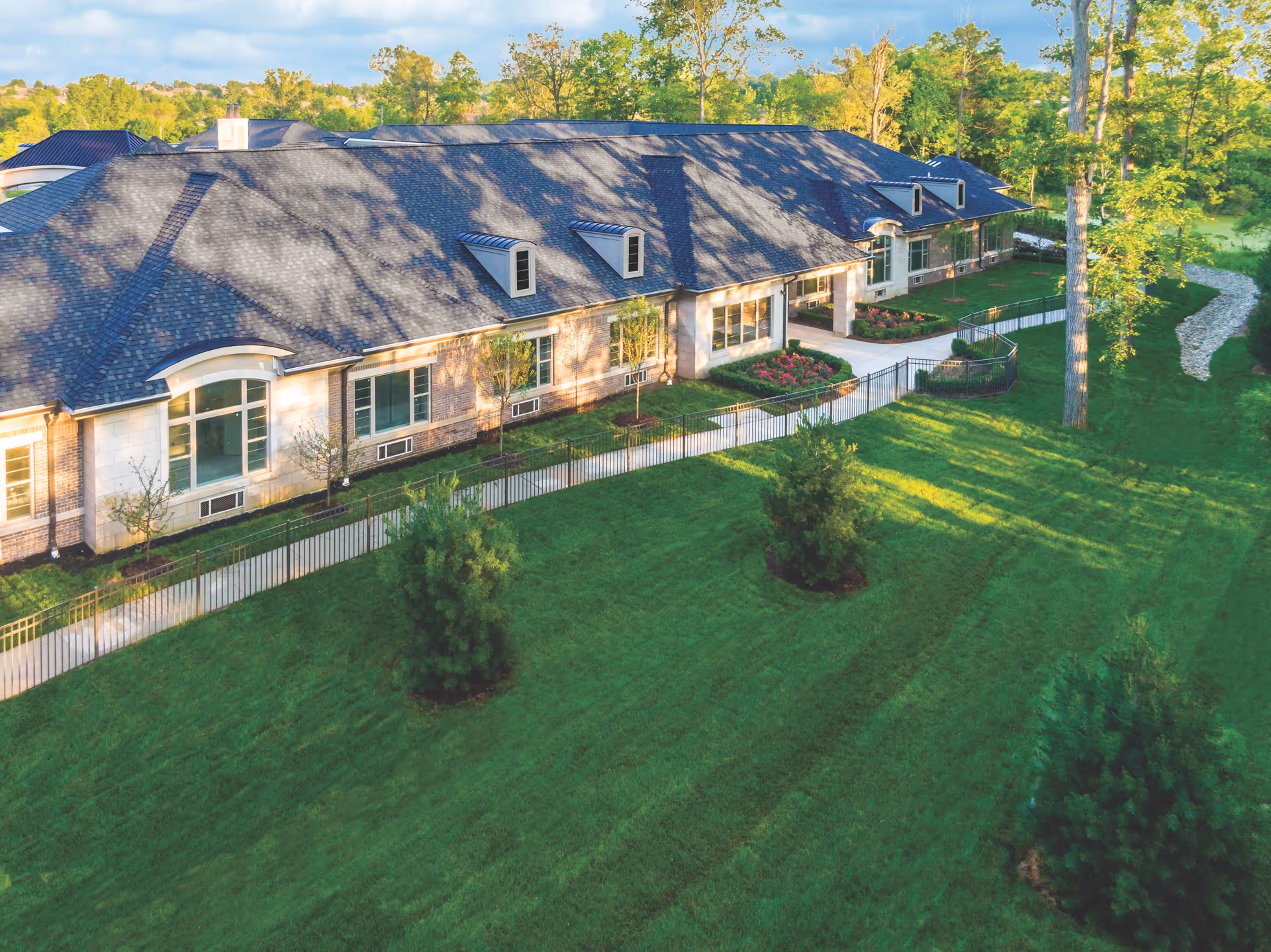 Aerial view of a long single-story building with large windows, a sidewalk, landscaped flower beds, and a green lawn surrounded by trees.