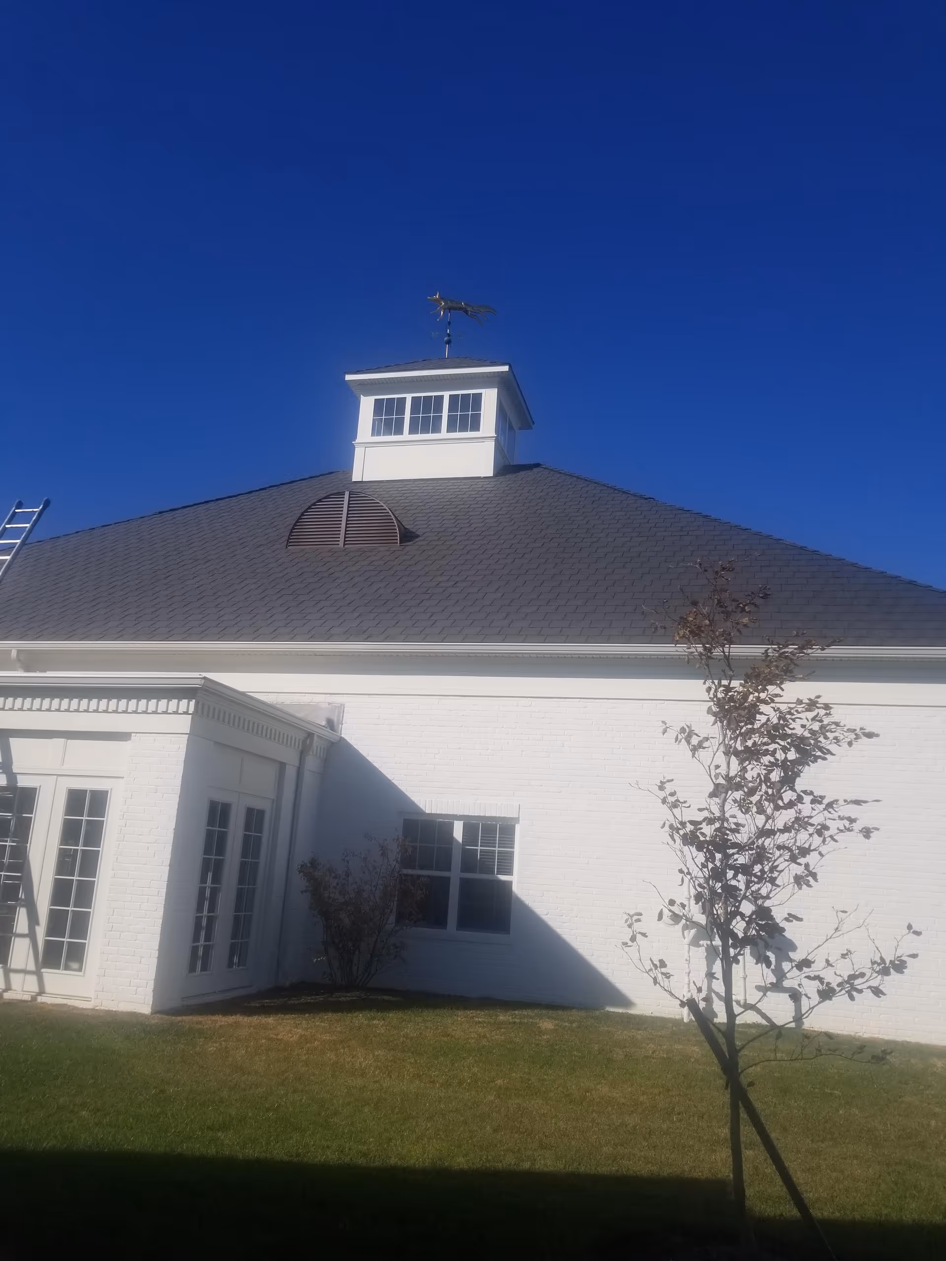 White brick senior living facility exterior with cupola, ladder, and small trees against a blue sky.