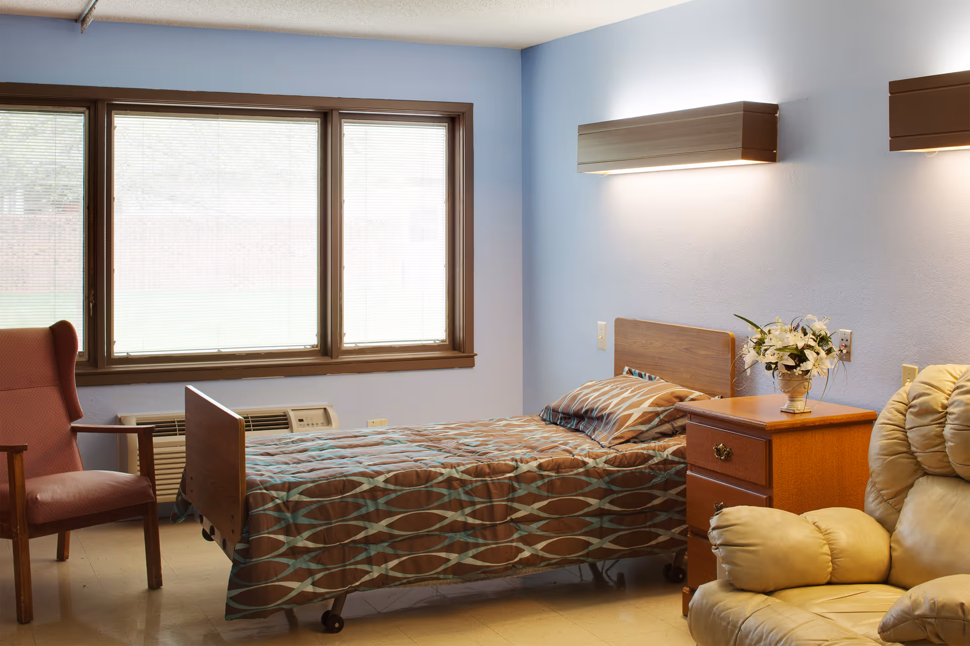 A single bed with a patterned brown and blue bedspread in a room with light blue walls. Next to the bed is a wooden nightstand with a vase of white flowers. There is a beige recliner chair in the foreground and a red armchair near a large window with blinds. The room has a simple, clean, and cozy appearance.