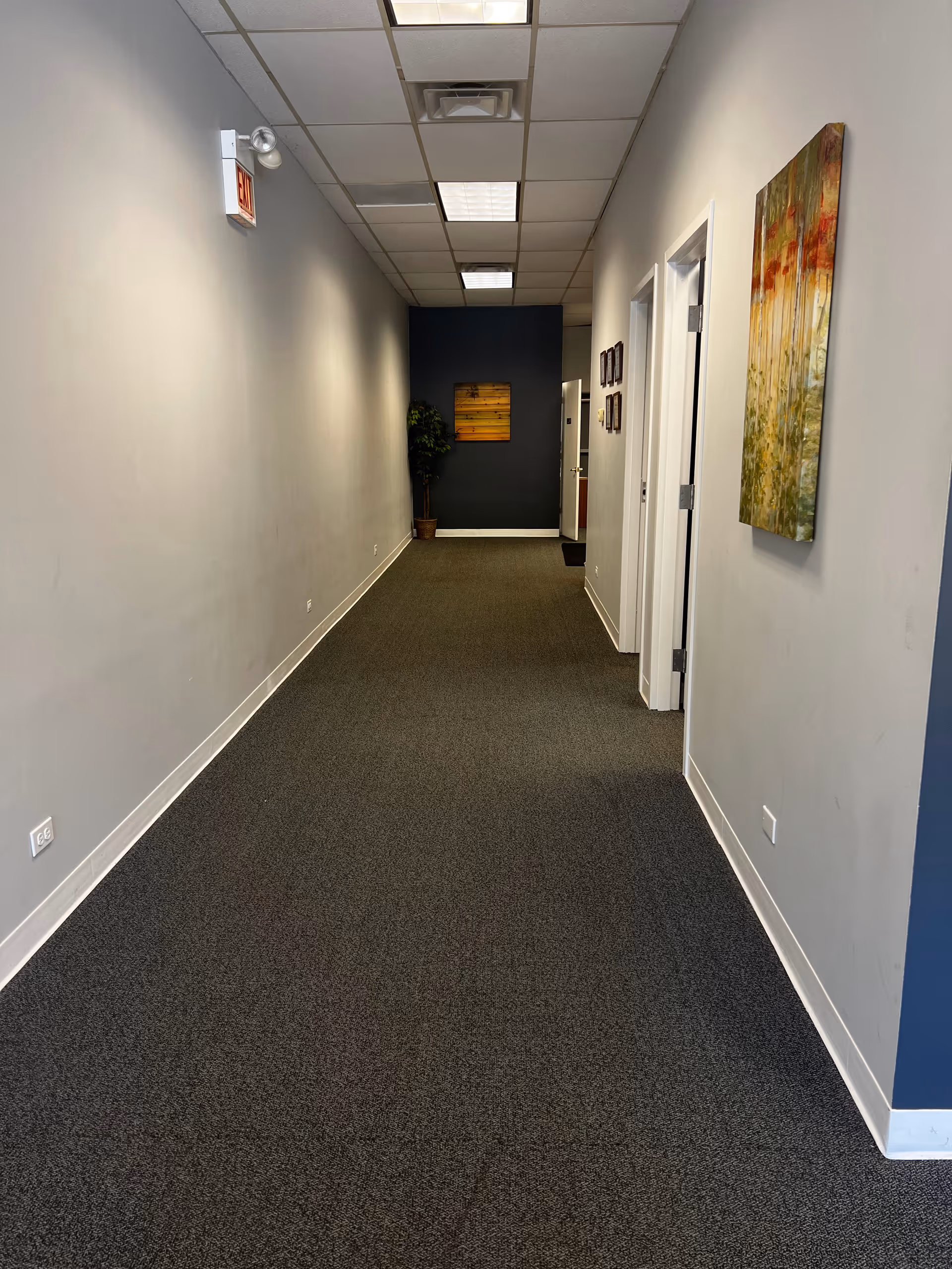 Long carpeted hallway in a senior facility with artwork and an exit sign.