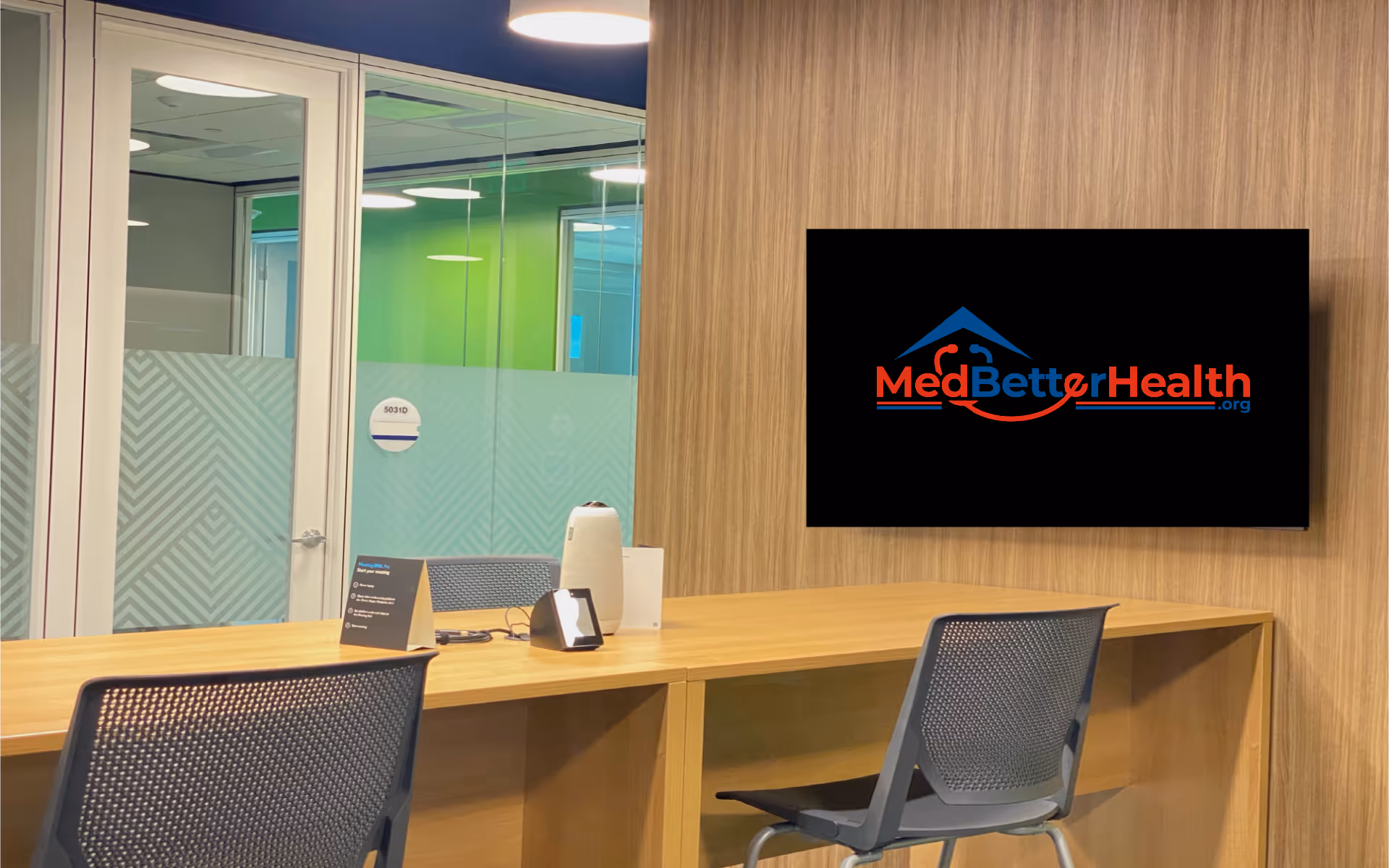 Reception area with wooden desk, two chairs, glass doors, and a wall-mounted MedBetterHealth logo on screen.
