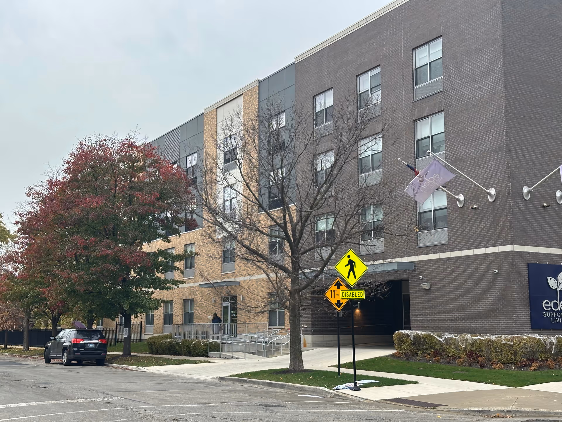 Exterior view of a multi-story senior living facility building with a mix of brick and panel siding. There are trees with autumn foliage along the sidewalk, a parked black SUV, and a pedestrian crossing sign with a disabled access sign below it. The building has flags mounted near the entrance and a sign that reads 'eden SUPPORTIVE LIVING'.