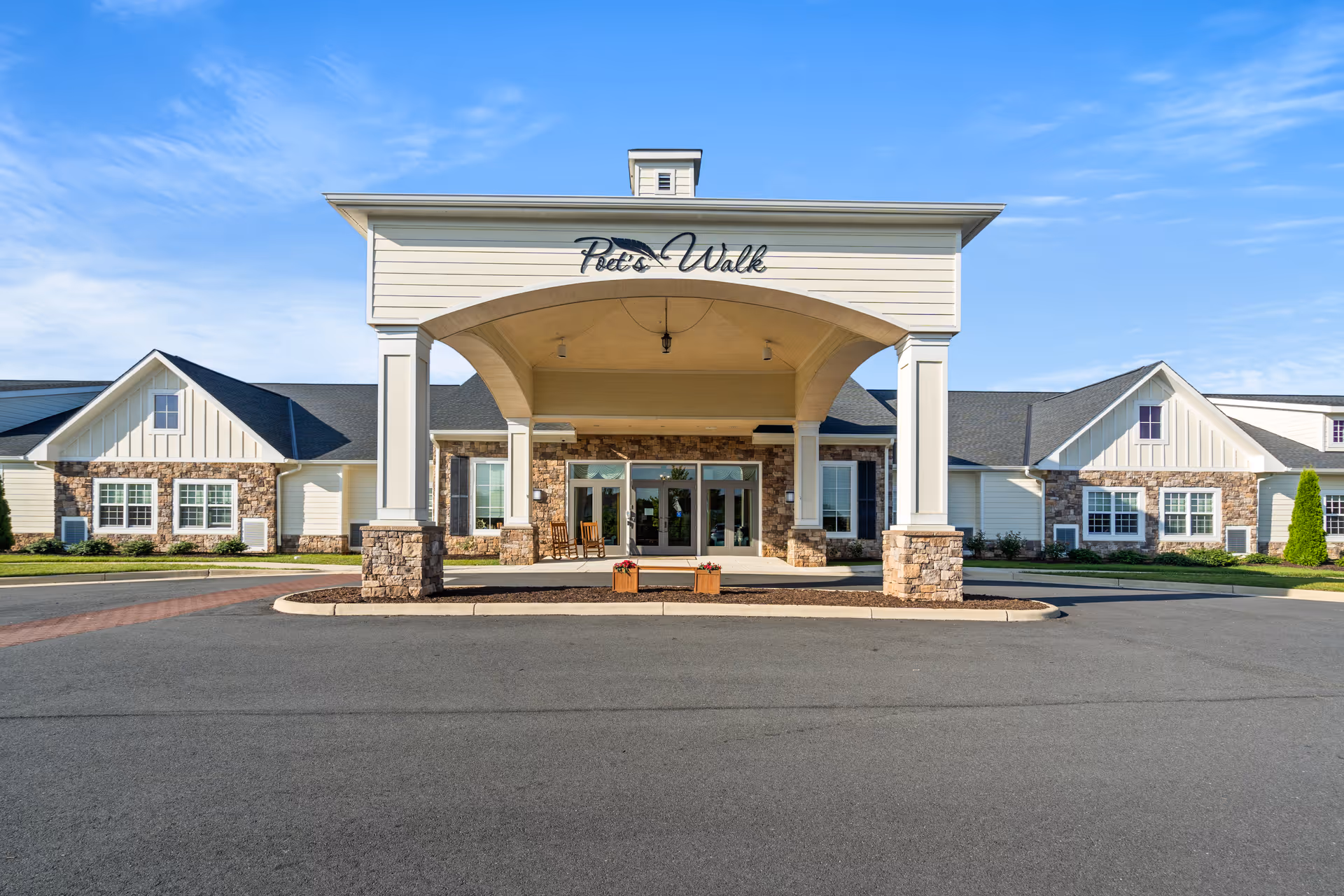 Front exterior view of Poet's Walk Leesburg, a memory care community, showing a large covered entrance with stone pillars, double glass doors, and a clear blue sky above.