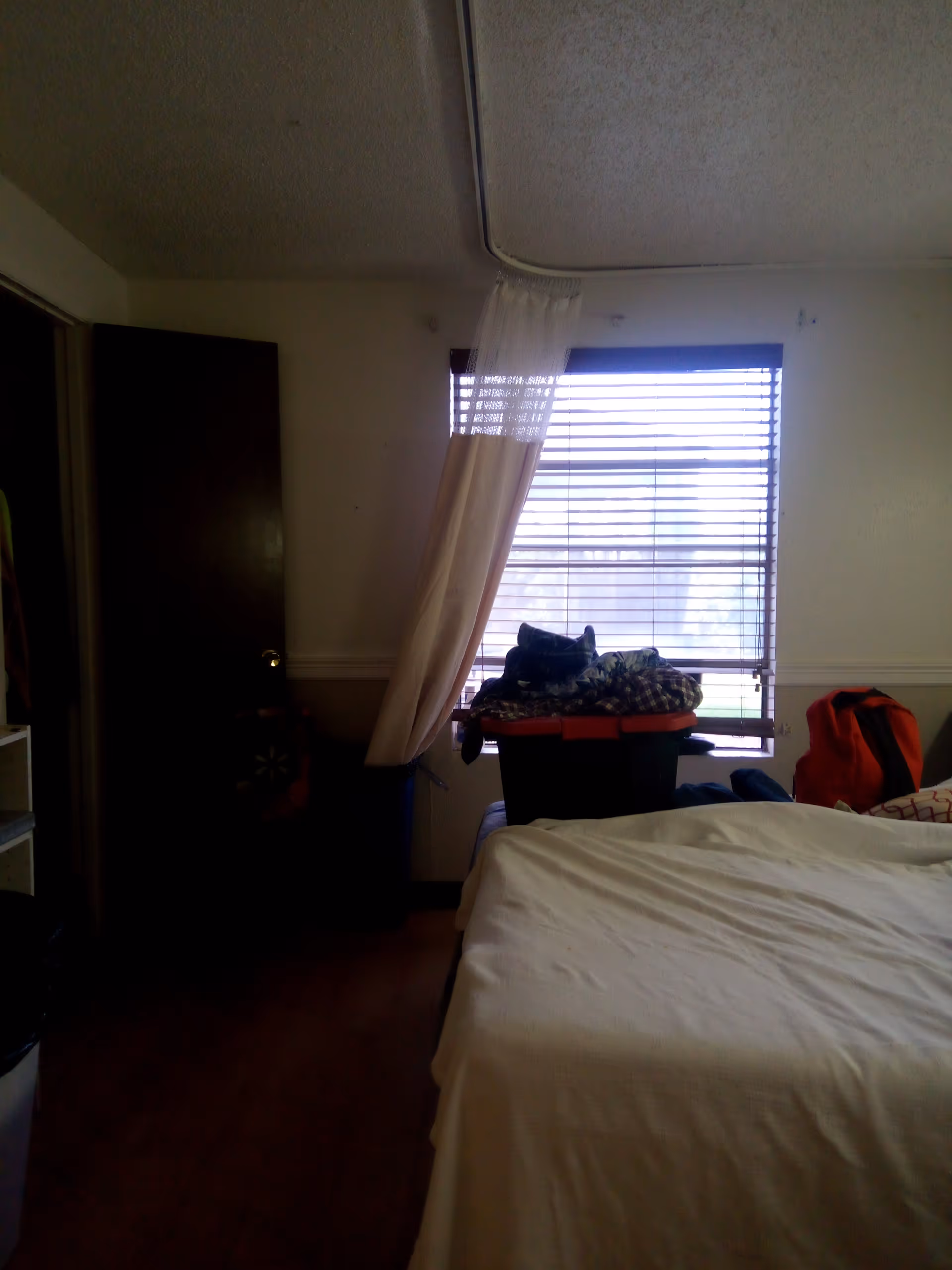 A bedroom with a bed, window blinds, and a pile of clothes near a laundry bin.