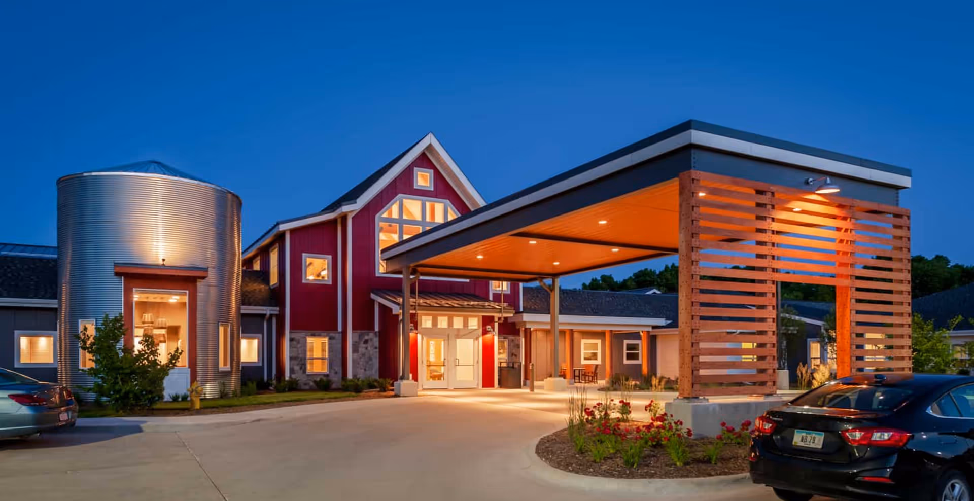 Front entrance of a red barn-style senior living community at dusk with a covered porte-cochère and parked cars.