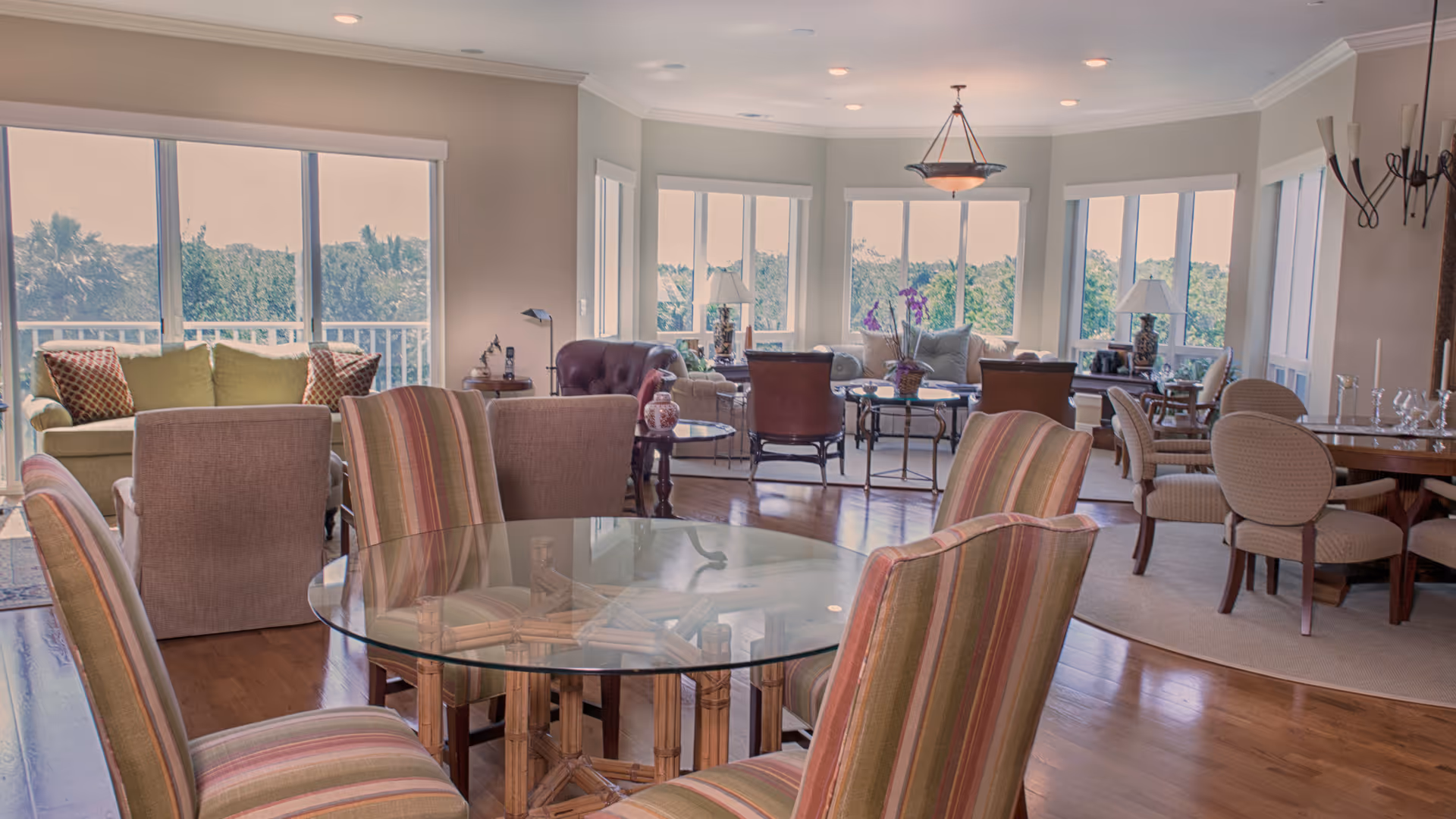 Spacious and well-lit common area with multiple seating arrangements including a glass-top round table with striped chairs in the foreground, several armchairs and sofas near large windows offering a view of greenery outside, and a dining table with upholstered chairs on a rug to the right.