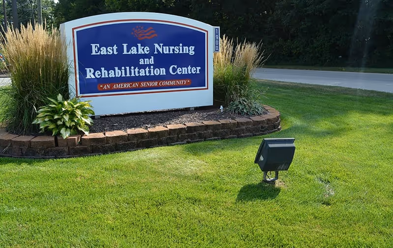 A large blue and white sign for East Lake Nursing and Rehabilitation Center surrounded by landscaping with green grass, plants, and ornamental grasses, located near a road.