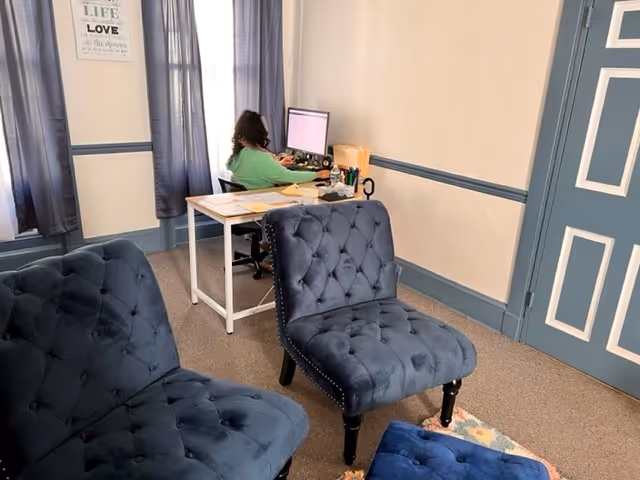 A desk area with a person at a computer, blue tufted chairs in a hallway-like room.