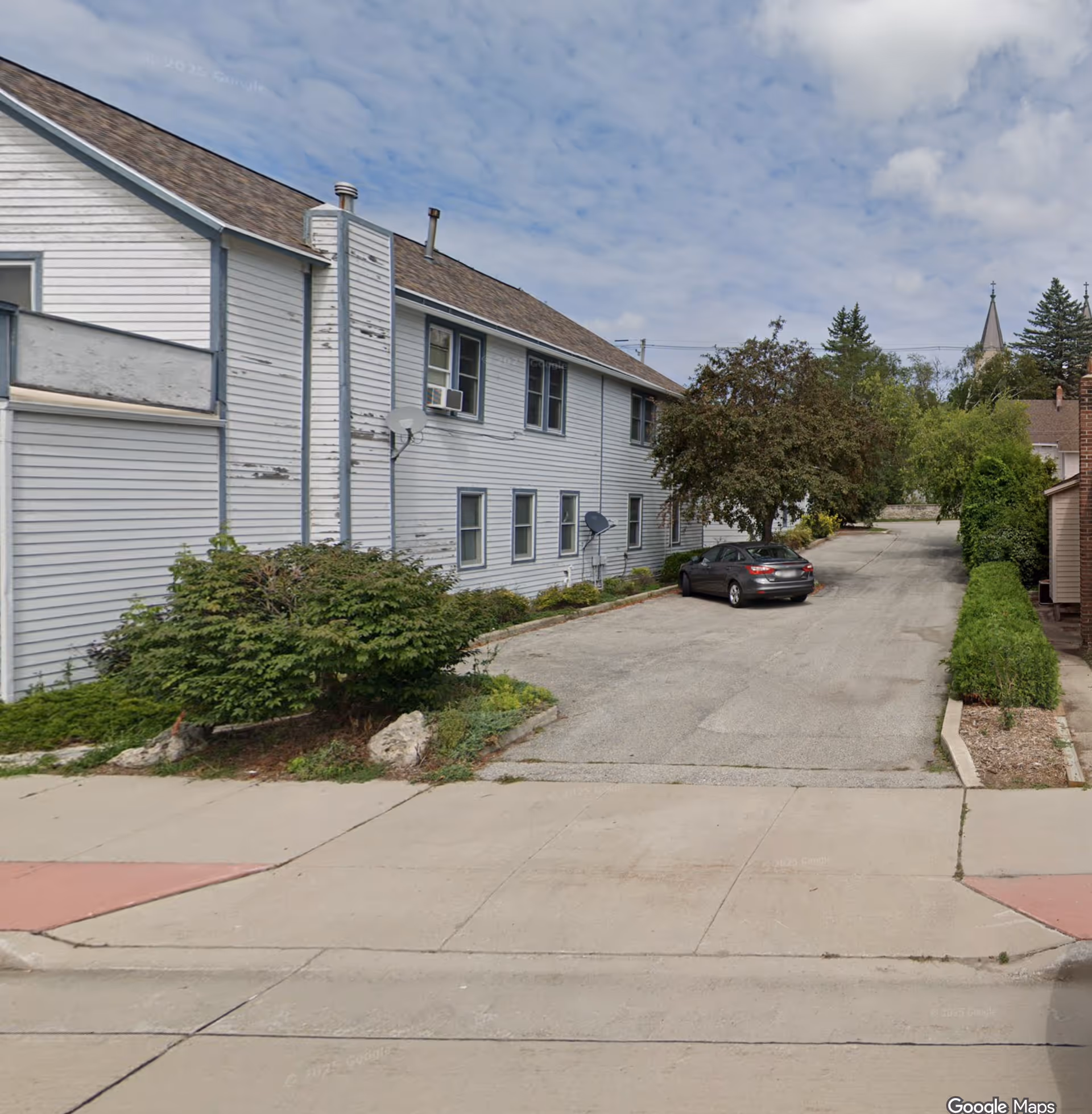 Long pale-blue building exterior with a driveway, parked car, and shrubs.