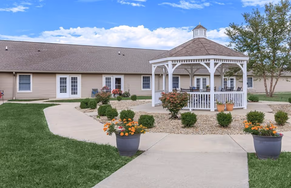 Outdoor courtyard of a senior living facility featuring a white gazebo, paved walkways, potted flowers, and a one-story building in the background.