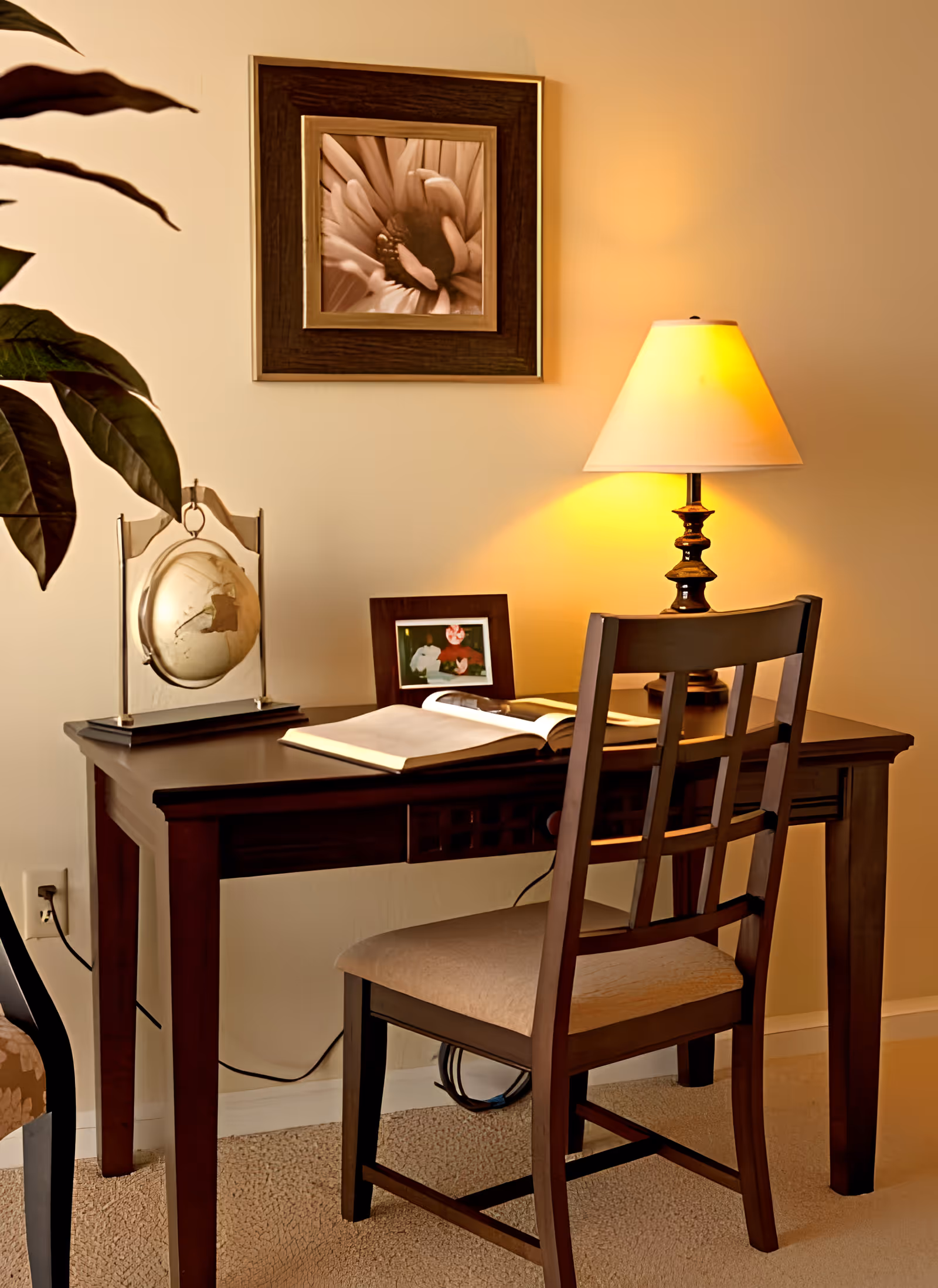 Wooden desk and chair in a warmly lit interior with a table lamp, open book, framed photo, decorative globe, and wall art.