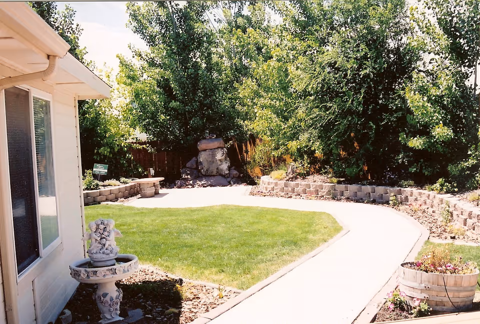 A sunny outdoor garden area with a curved concrete pathway, green grass, a stone water feature in the background, and a decorative birdbath near the side of a light-colored building. There are trees and plants along the edges of the garden.