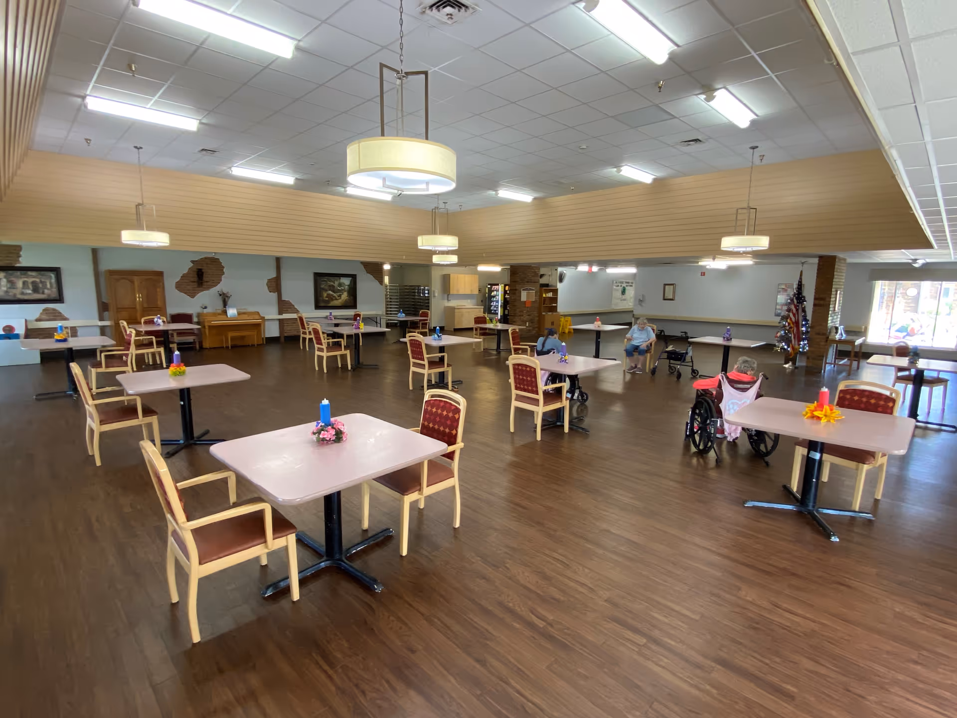 Large communal dining hall with several tables, chairs, and a few residents seated.