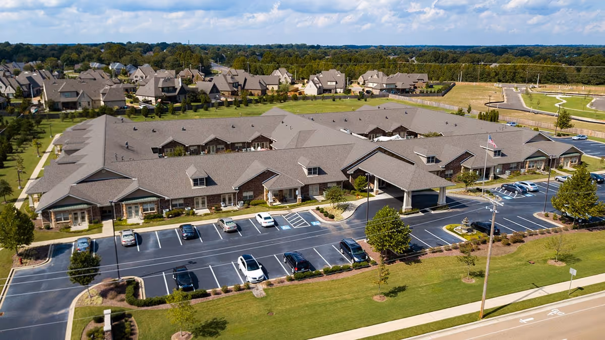 Aerial view of Hearthside Senior Living facility showing a large single-story building with a covered entrance, surrounded by a parking lot with several cars parked. The building is set in a suburban area with green lawns, trees, and residential houses in the background under a partly cloudy sky.