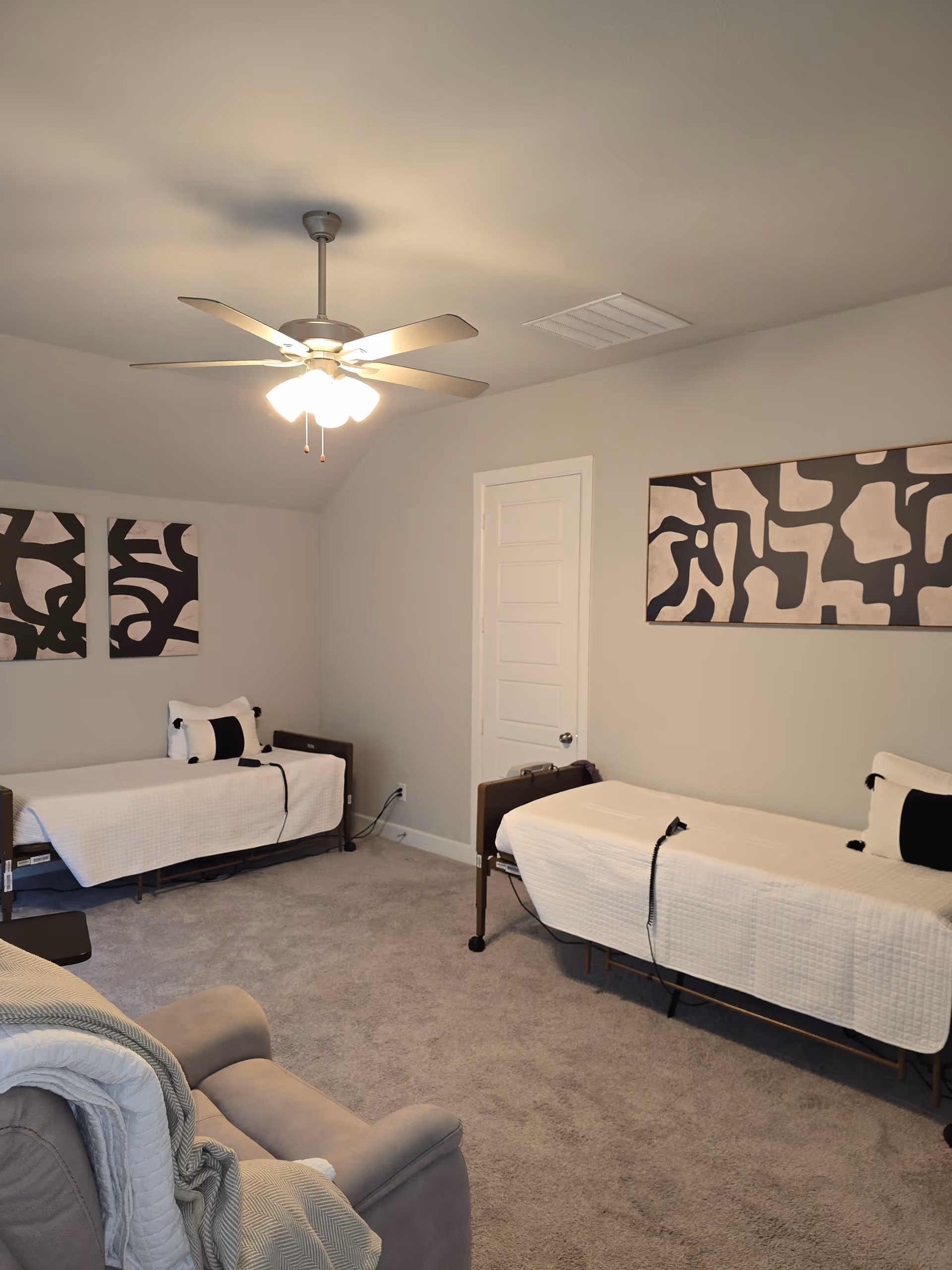Two single beds with white bedding in a gray-walled senior living bedroom with a ceiling fan.
