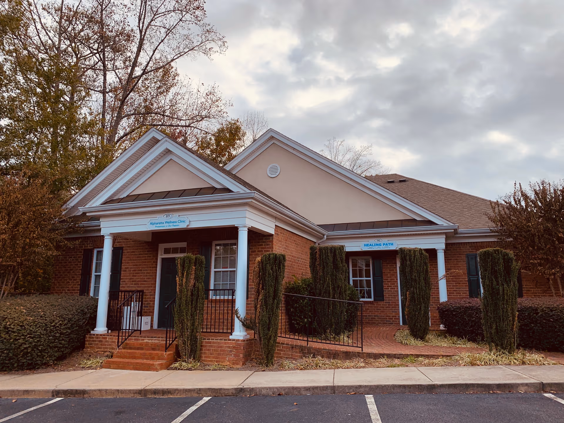 Brick facility with white columns, a small front porch, signage, and tall hedges along the entrance.