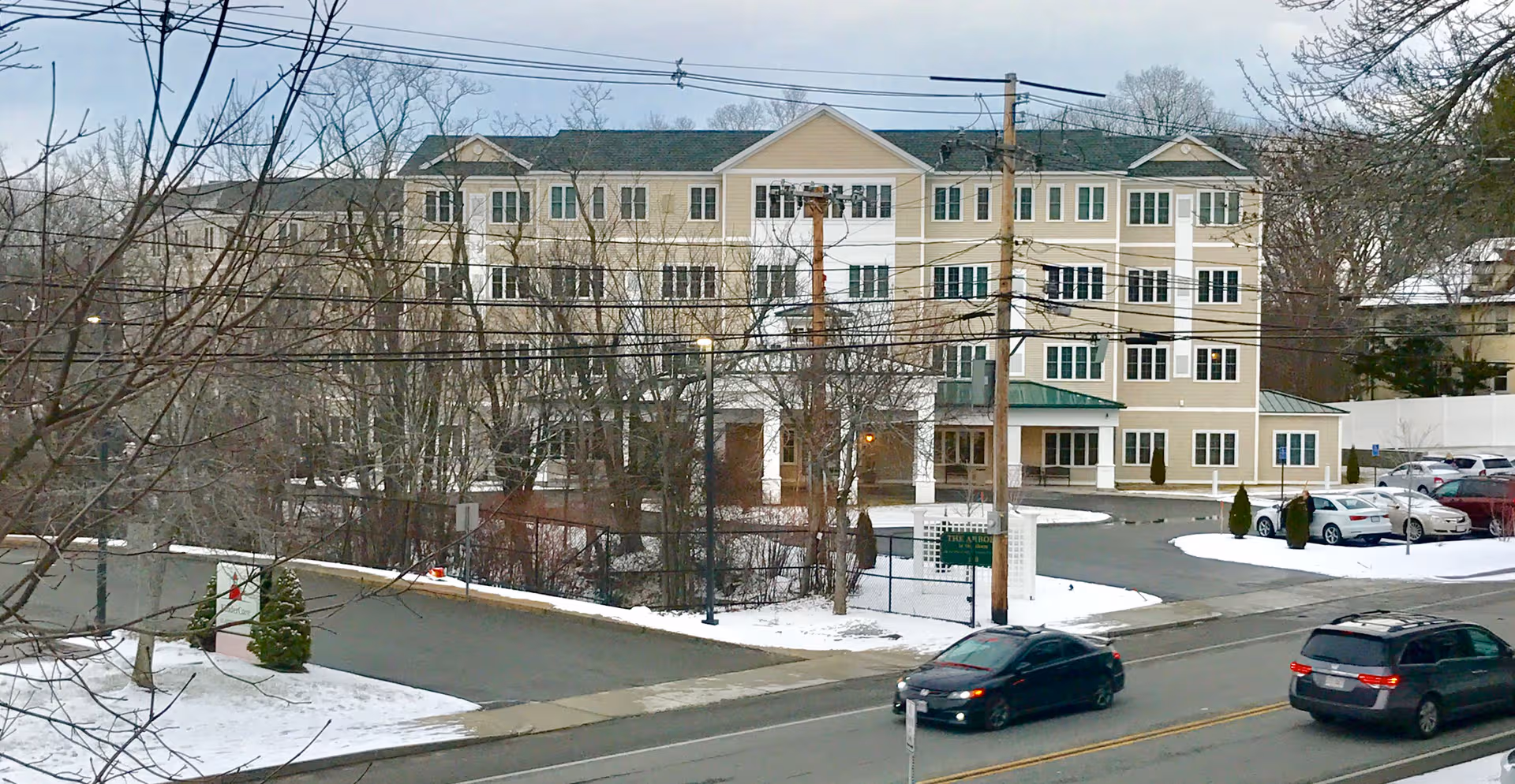 Three-story beige senior living building viewed from across a snowy street with parked cars.