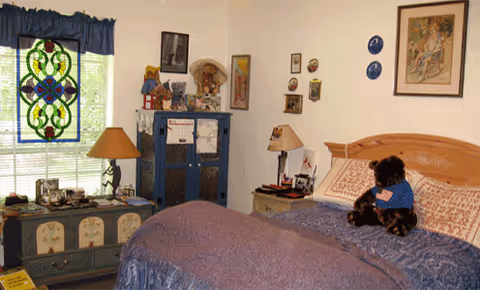 Cozy bedroom with wooden bed, quilt, stained glass window, dresser, and a person with a dog.