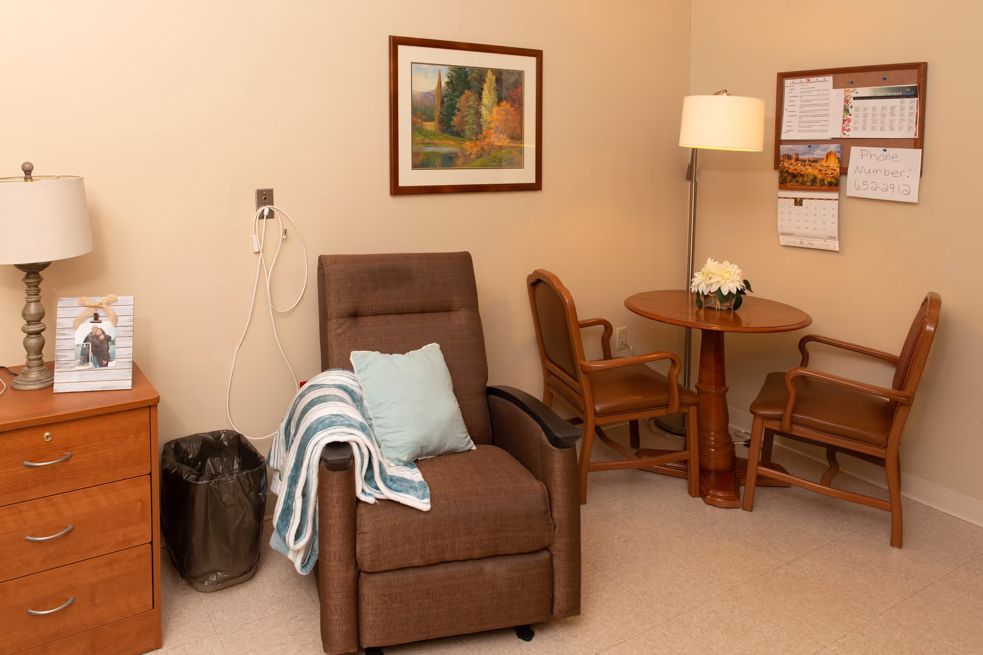 A cozy interior sitting area with a brown recliner draped with a blanket and pillow, a wooden side table and lamp, and a small round table with two chairs against a bulletin board.