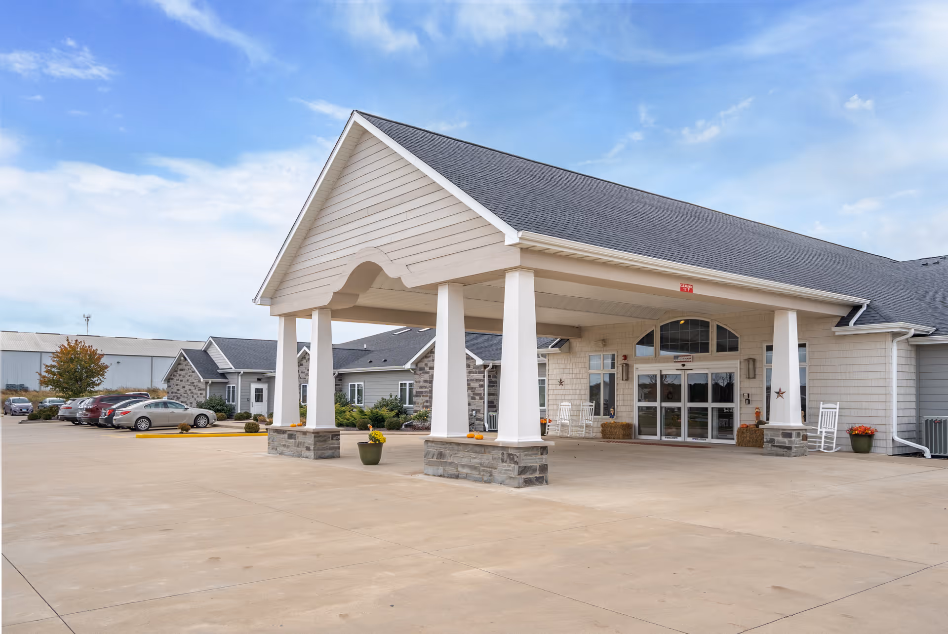 Covered entrance and drop-off canopy of a single-story memory care facility with columns, rocking chairs and a parking lot.