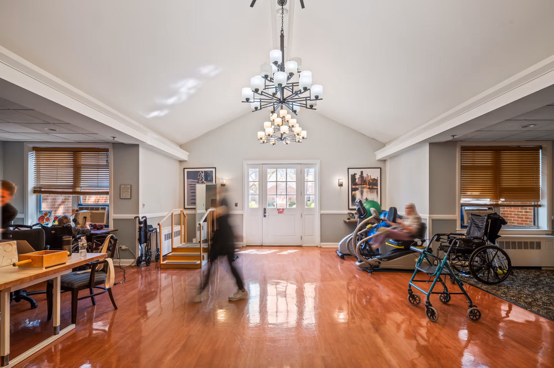 Bright senior living common area with exercise equipment, wheelchairs, and activity tables.