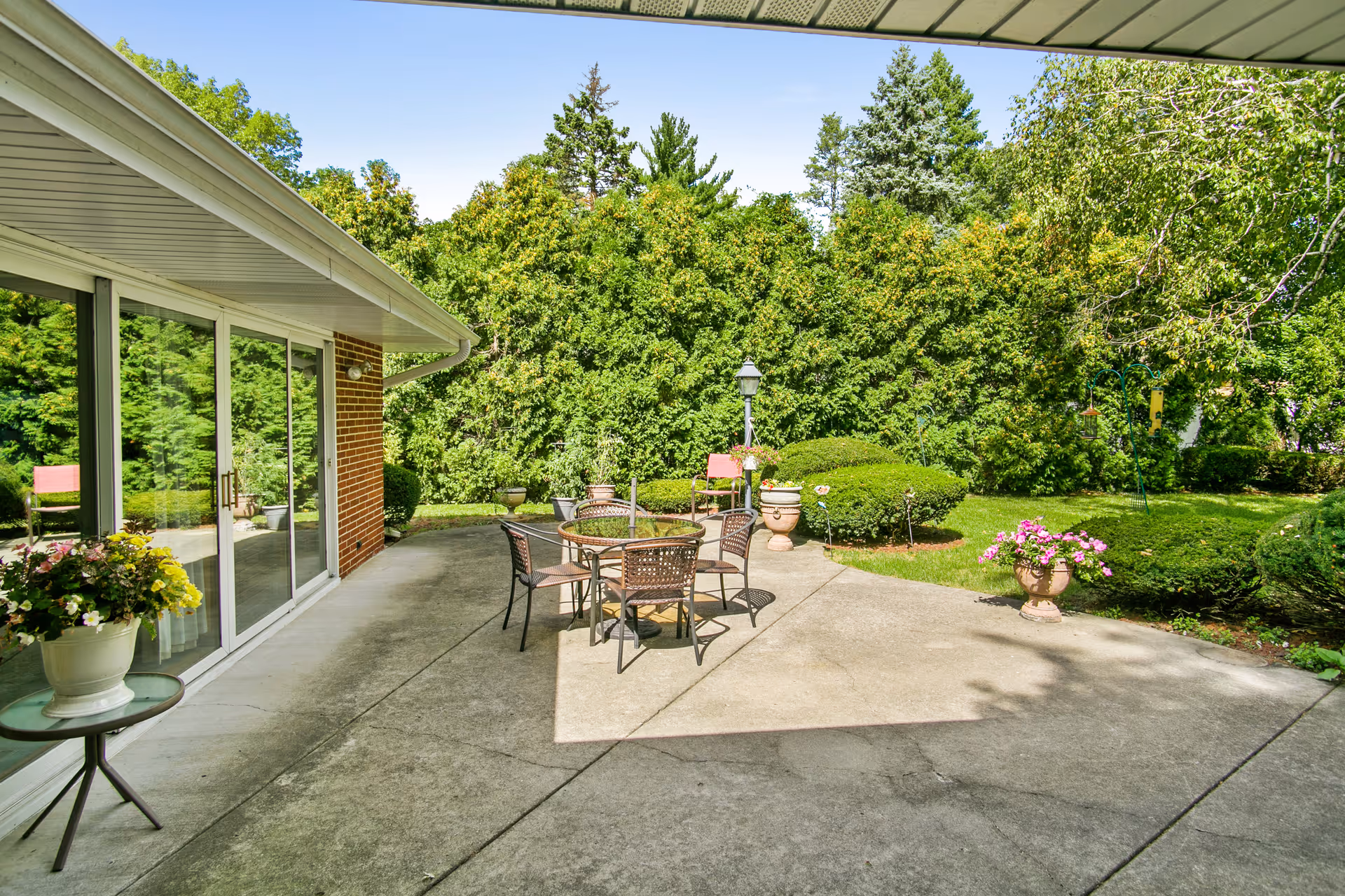 Sunlit concrete patio with a round table and chairs, potted flowers, sliding glass doors, and a hedge-lined backyard.