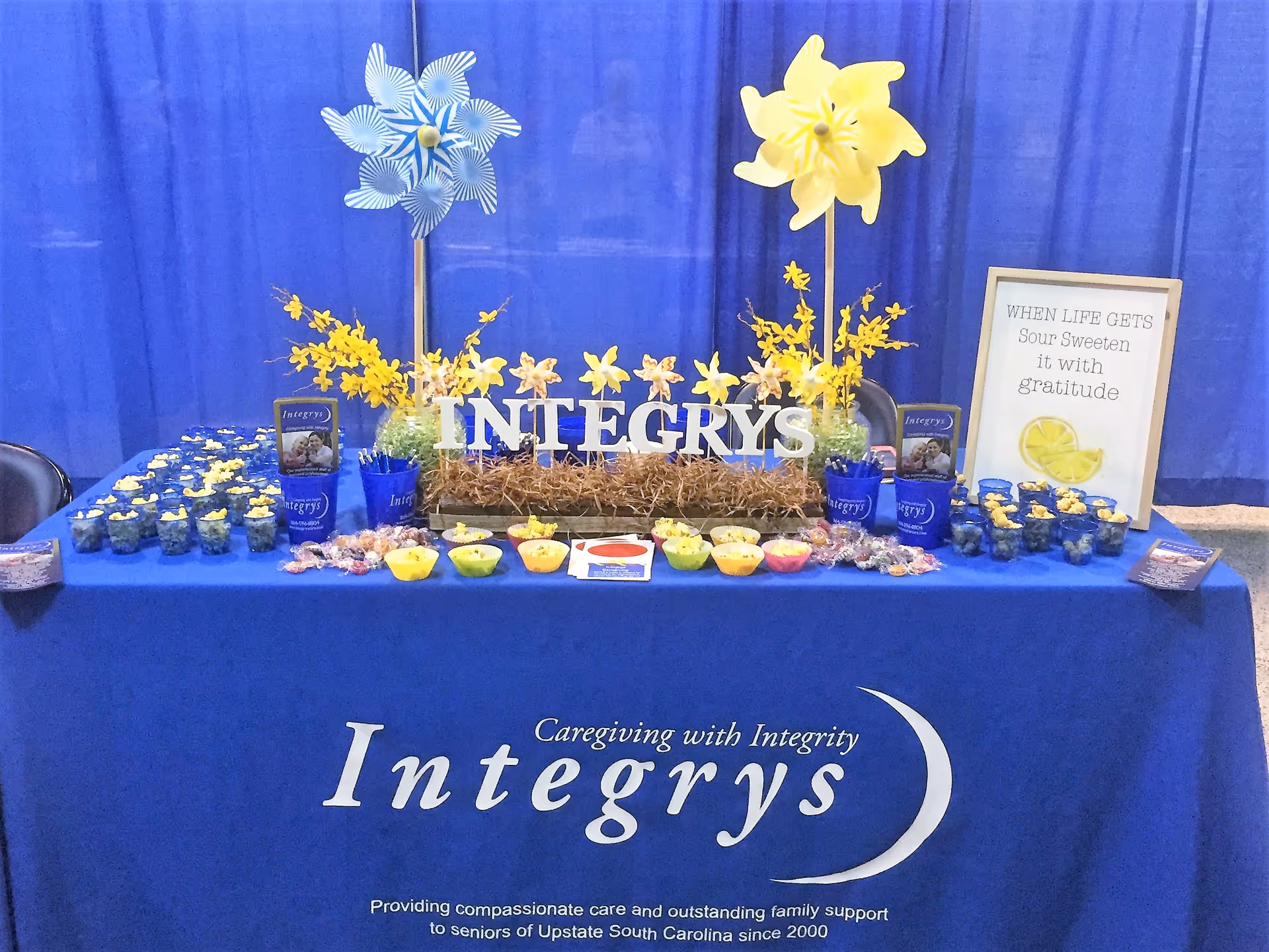 Blue promotional booth table with Integrys branding, yellow flowers, cupcakes, and informational materials.
