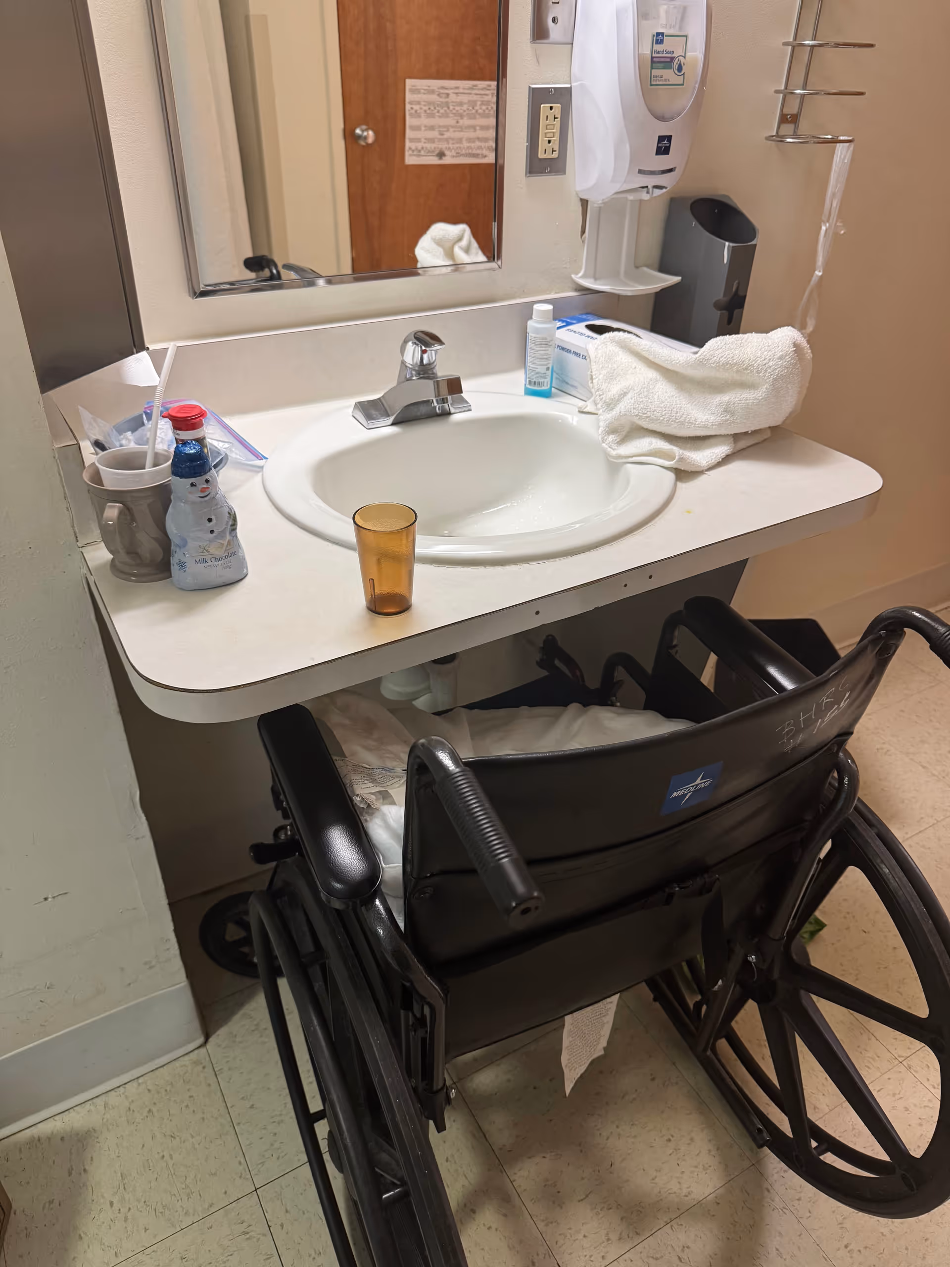 Wheelchair at a bathroom sink with mirror and toiletries.