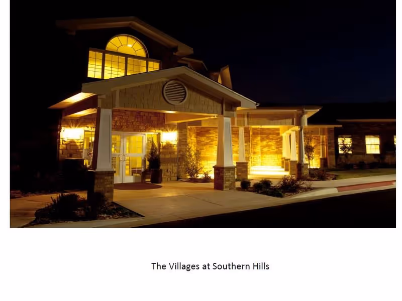 Night view of the entrance to a building with warm yellow lighting illuminating the porch area, stone and brick exterior walls, and large windows. The building has a covered entryway supported by white columns.