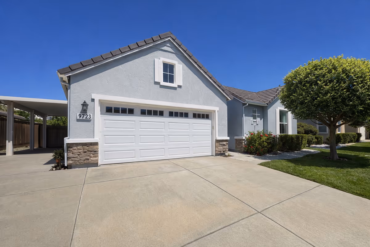 Light gray, single-family home with a two-car garage, driveway, manicured lawn, and tree.