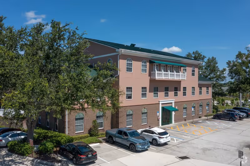 Two-story senior living facility with pink brick facade, green roof, and parking in front.