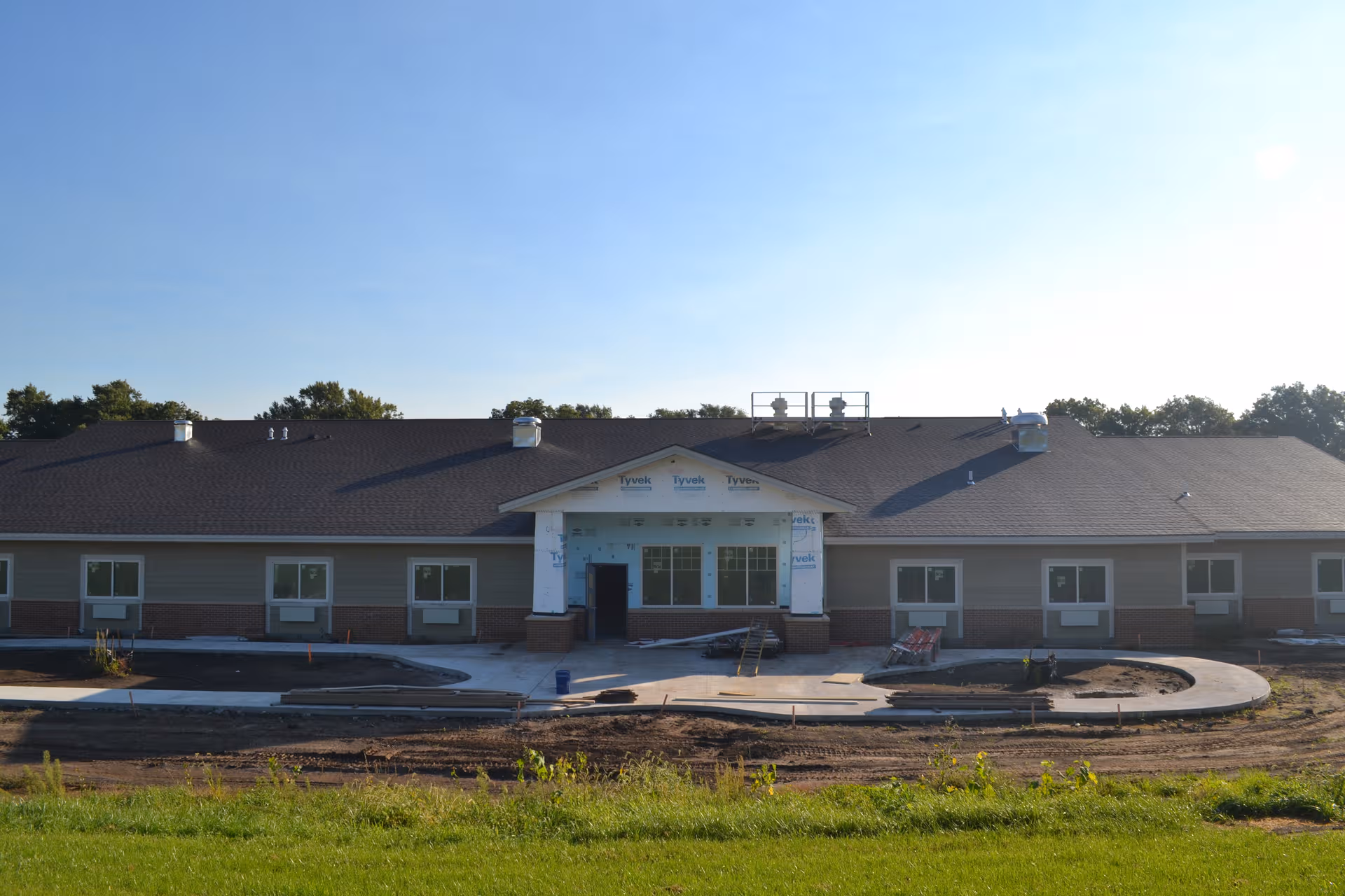 Long, single-story facility under construction with Tyvek-wrapped entrance and new sidewalk.