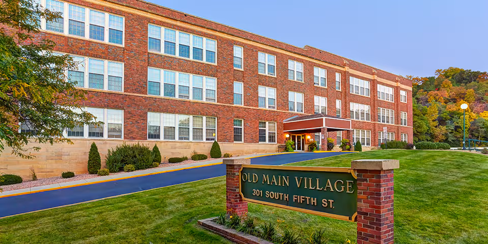 Exterior view of Old Main Village, a senior living facility, with brick building and green lawn.