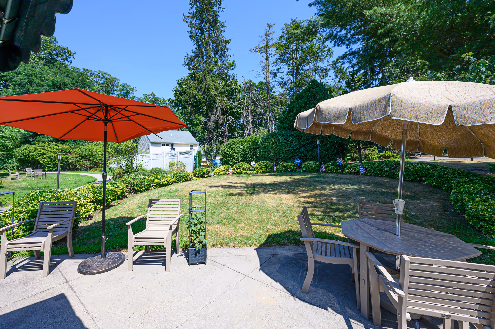 Outdoor patio area with beige chairs and tables under large umbrellas, one red and one beige, surrounded by green bushes and trees under a clear blue sky.