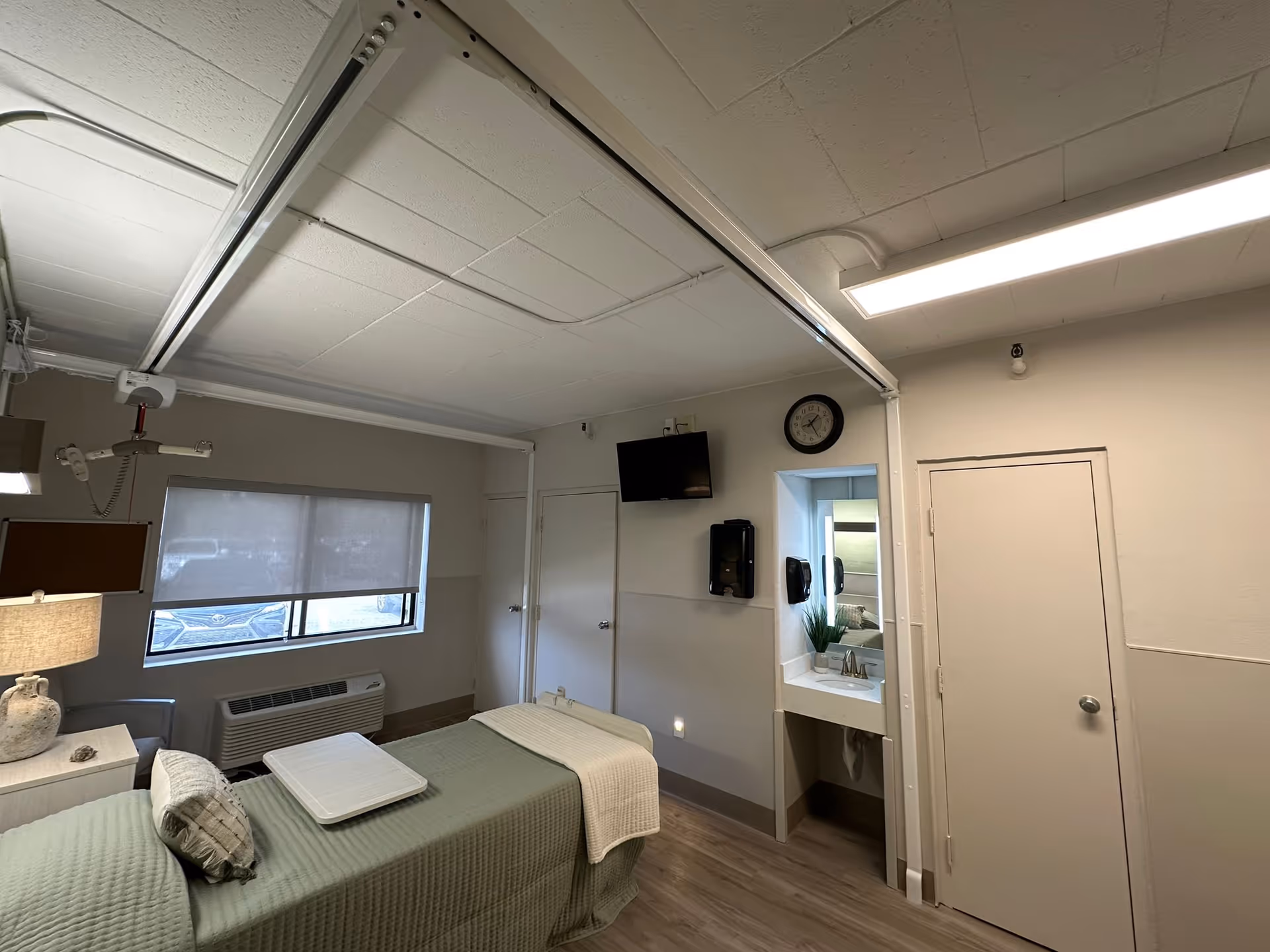 A nursing facility room with a single bed covered in a green quilt and a white blanket, a small bedside table with a lamp, a window with a roller shade, a wall-mounted TV, a clock, a small sink with a mirror above it, and two closed doors. The room has light-colored walls and wood flooring.